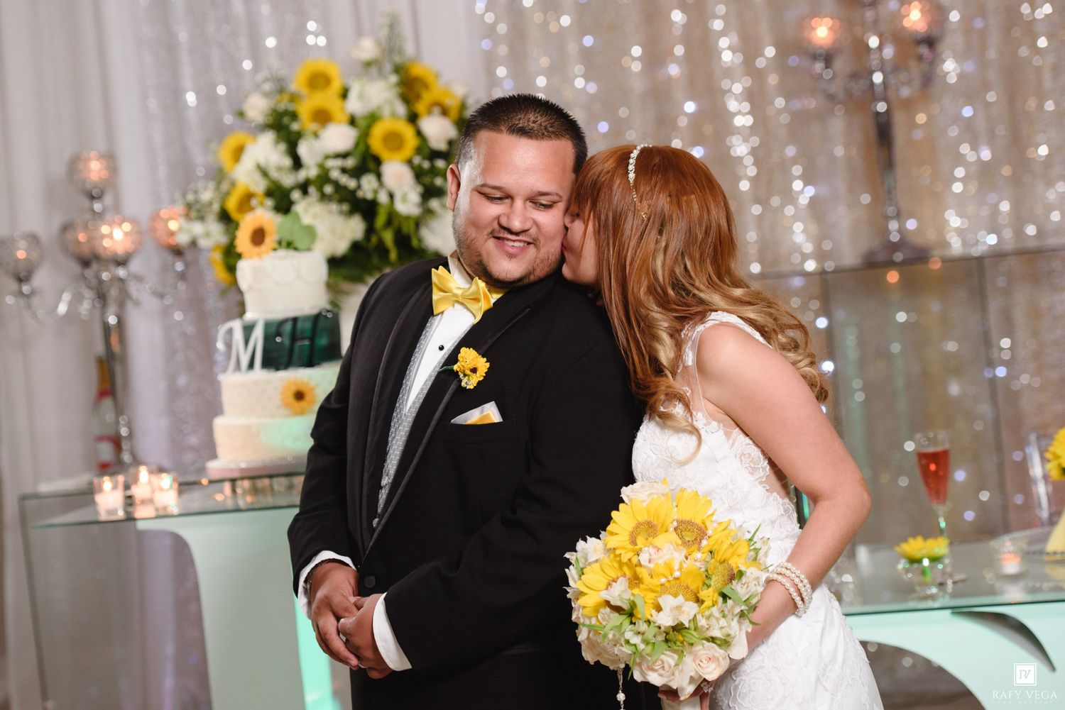 Bride kissing groom on cheek at wedding reception, groom in tuxedo with yellow bow tie, bride holding sunflower bouquet.