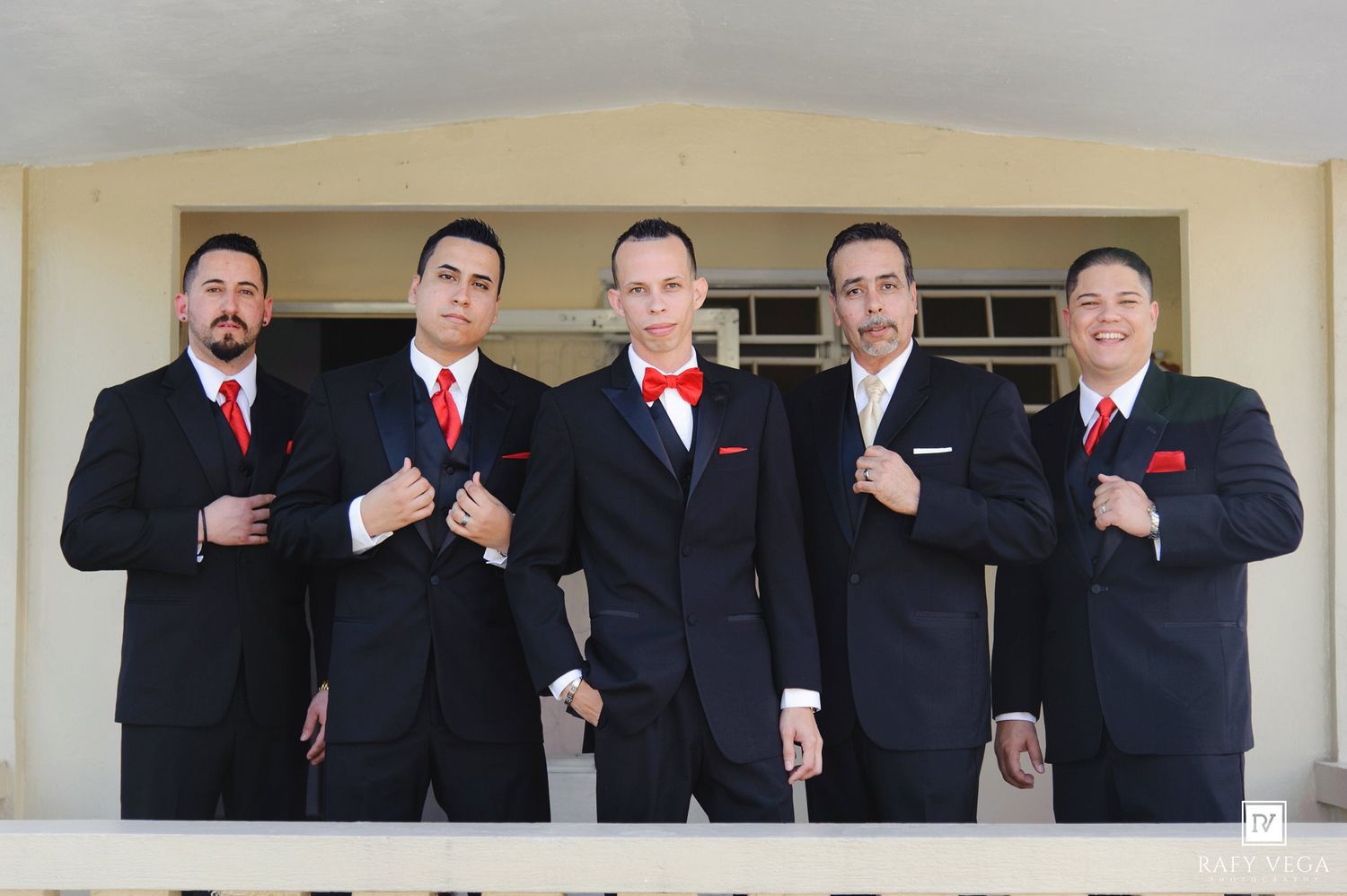 Five groomsmen in black suits with red ties and pocket squares pose confidently on a balcony for a wedding photo.