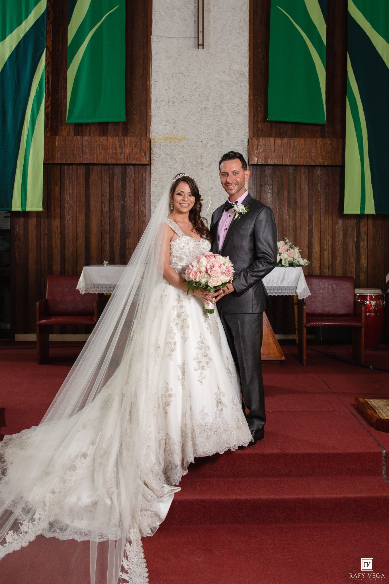 Bride and groom posing together at church altar, bride wearing lace ball gown with long veil, groom in gray suit.