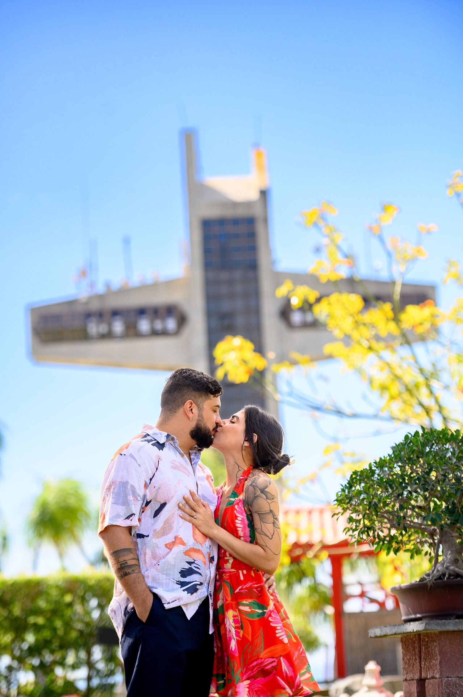 A couple shares a romantic kiss outdoors near a modern building and yellow flowers on a sunny day.