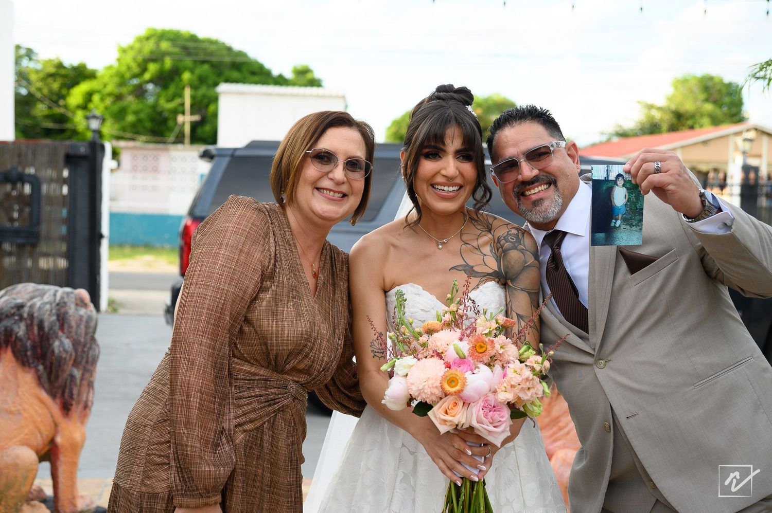 A smiling bride in a white gown holds a peach and pink bouquet while posing with a woman in a brown dress and a man in a gray suit outdoors.