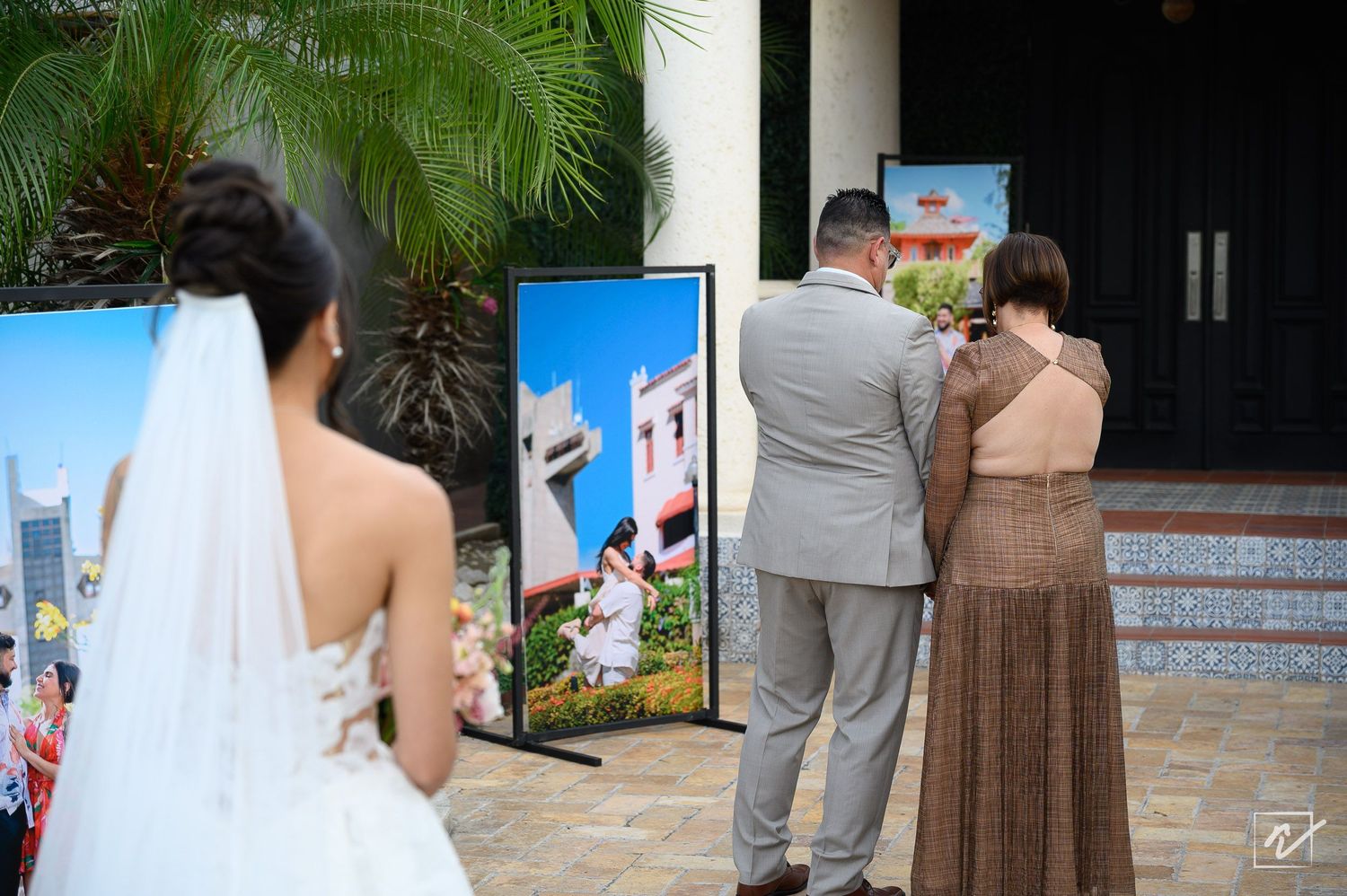 A bride in a white wedding gown and veil views framed photo displays outdoors as two guests in formal attire stand nearby.