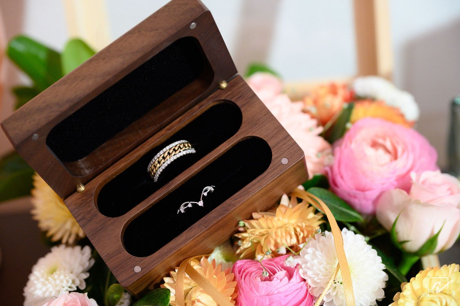 Wooden ring box with two wedding bands resting inside, surrounded by a bouquet of pink, white and orange flowers.