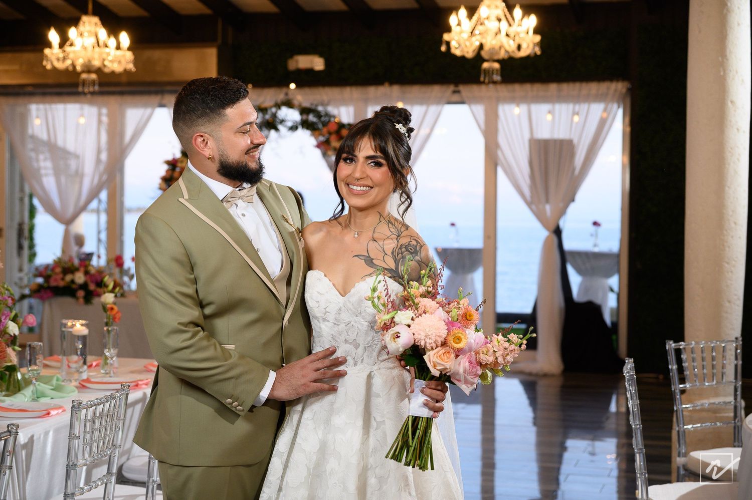 Smiling bride in a white gown holding a pink floral bouquet embraces her groom in an olive suit inside an elegant ballroom with chandeliers.