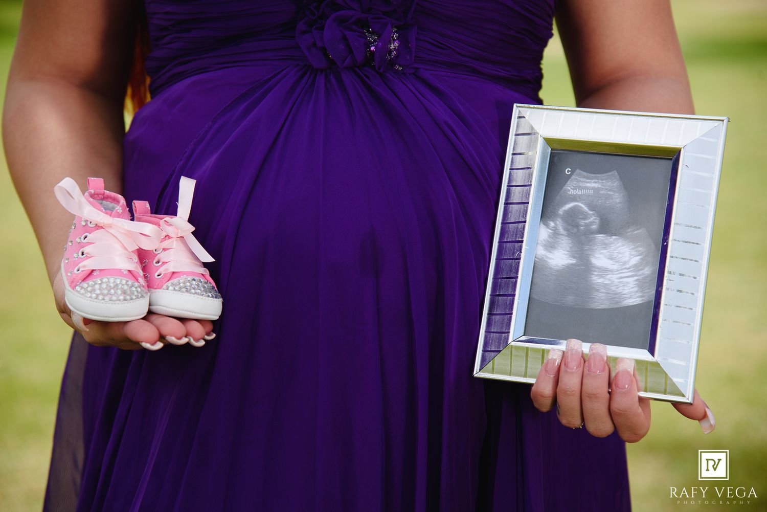 Pregnant woman in purple dress holding pink baby shoes and ultrasound photo outdoors.