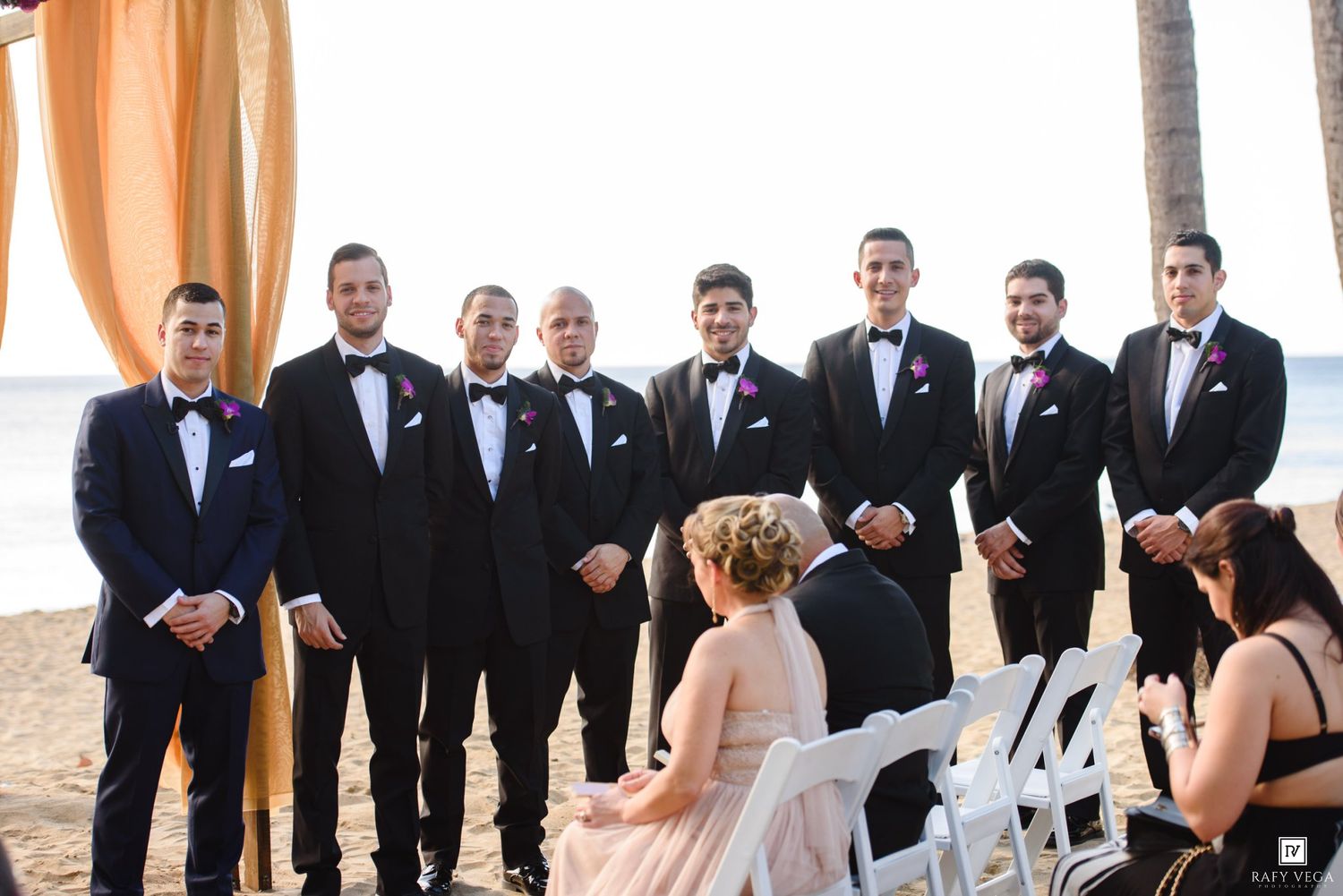 Groomsmen in black tuxedos stand on a beach during an outdoor wedding ceremony with white chairs and draped fabric.