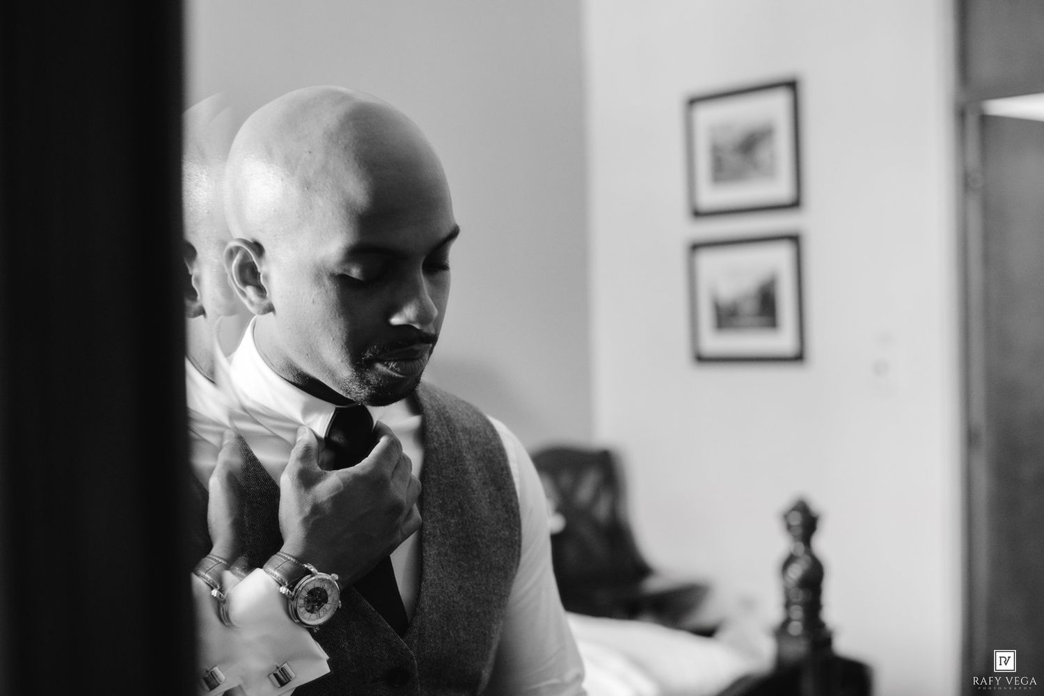 Black and white photo of a bald man adjusting his bow tie while getting dressed in formal attire.