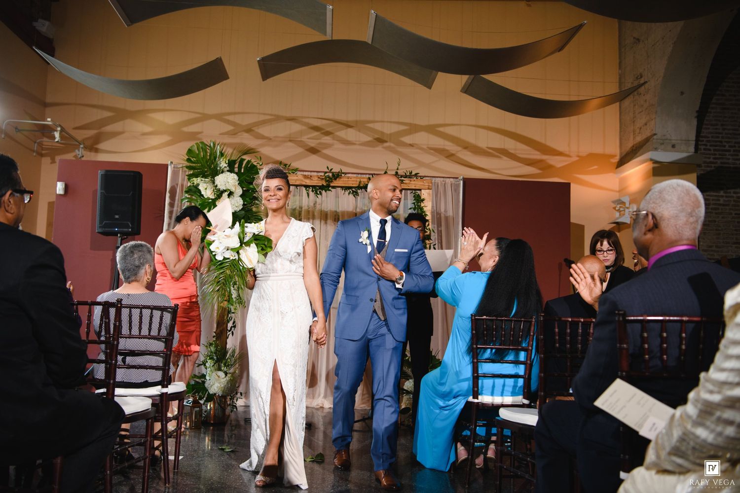 Newlywed couple walks down the aisle hand-in-hand as guests applaud at an elegant indoor wedding ceremony.