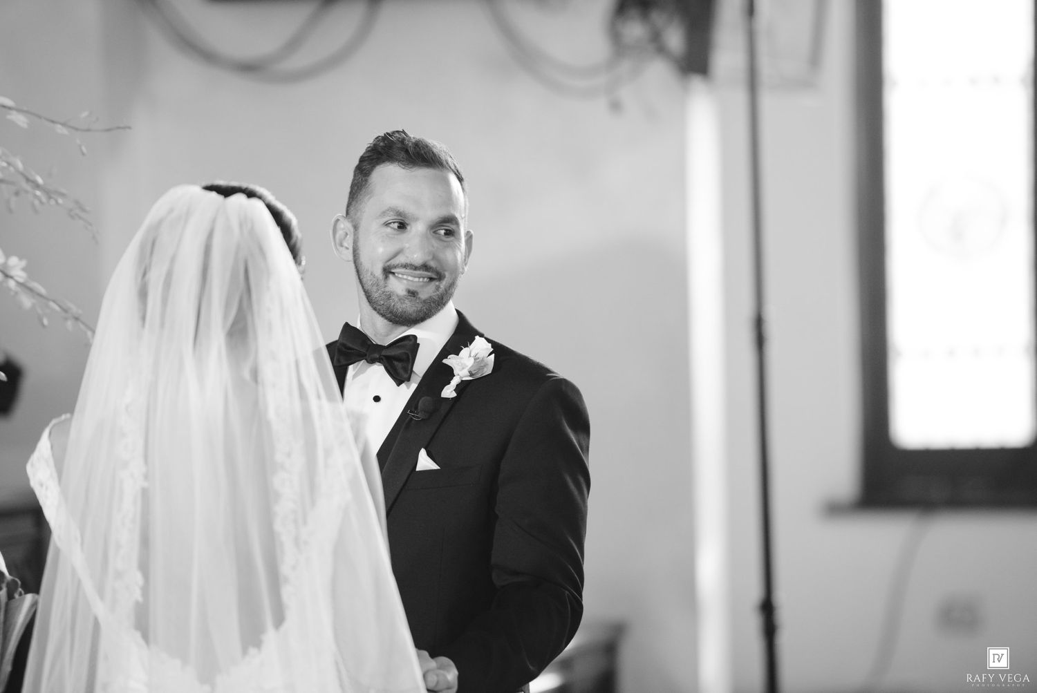 Black and white photo of a smiling groom in a tuxedo looking at his bride during a wedding ceremony.