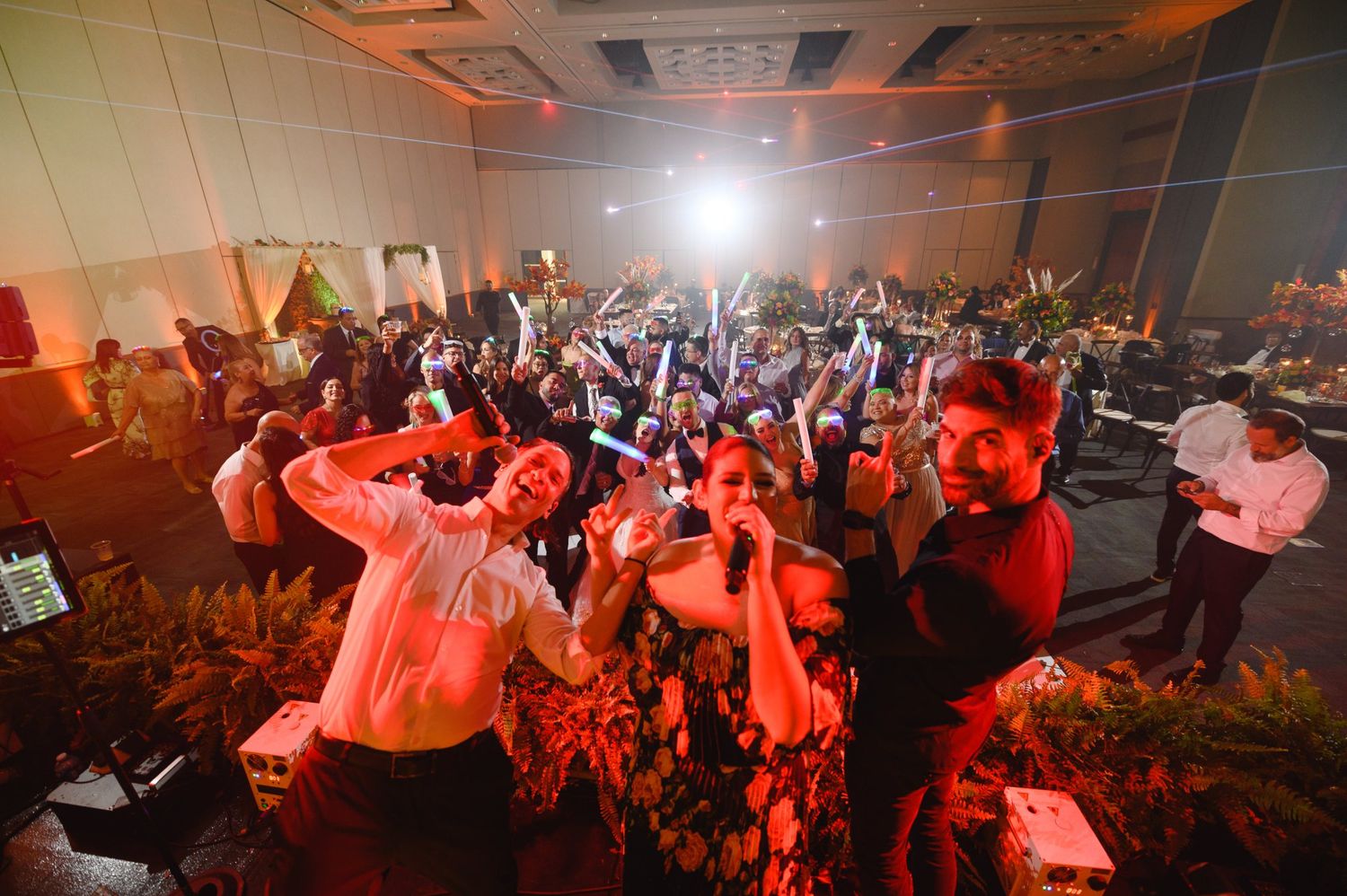 Energetic party crowd celebrating at an indoor event with colorful lighting and guests raising hands on the dance floor.