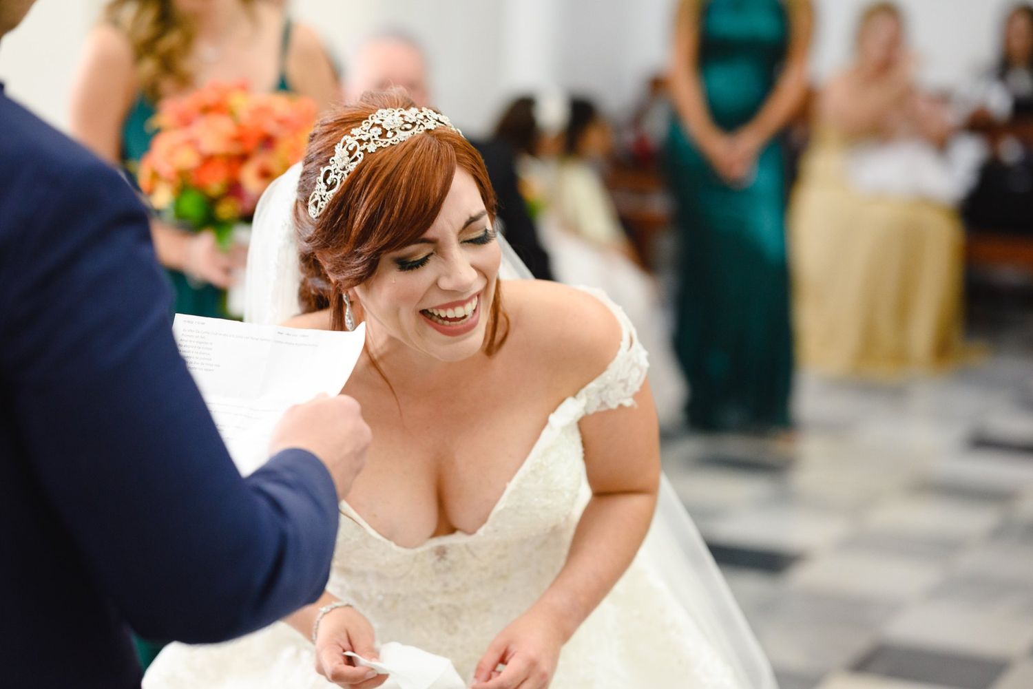 Smiling bride in white lace gown and gold tiara during wedding ceremony with bridesmaids in green dresses.