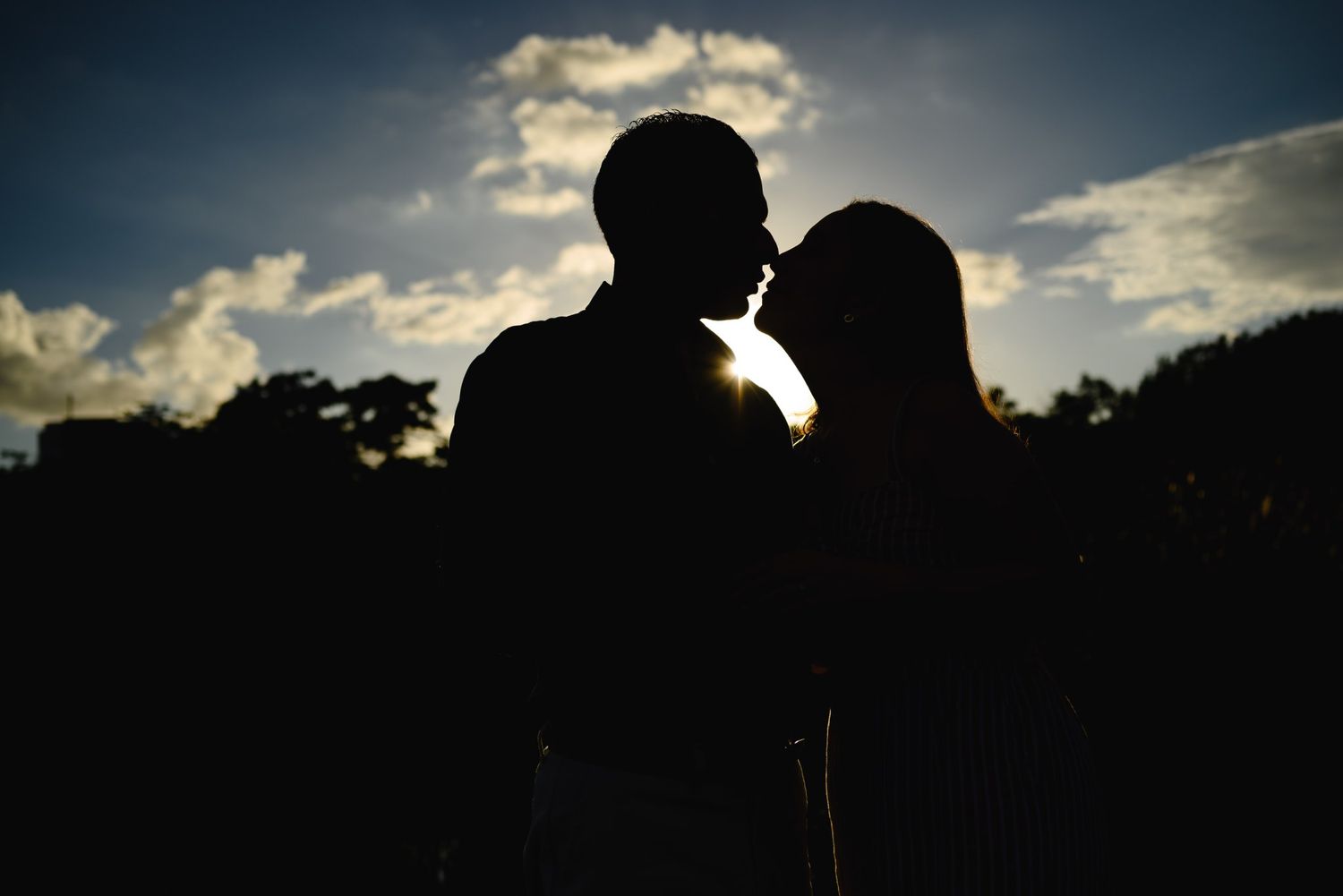 Romantic silhouette of a couple almost kissing against a dramatic sunset sky with clouds and trees.