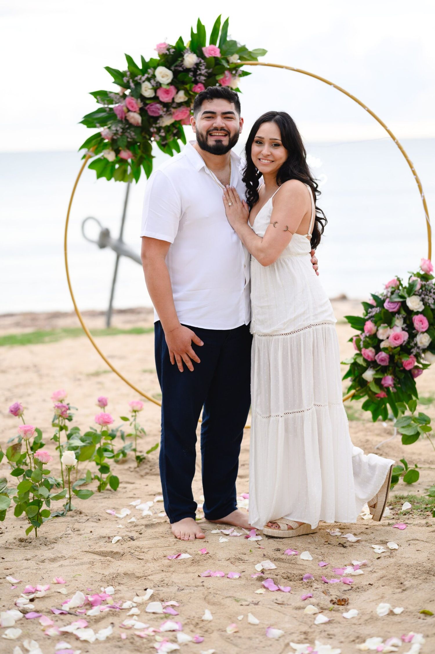 Couple poses under floral hoop arch on beach with pink roses and petals scattered on sand.