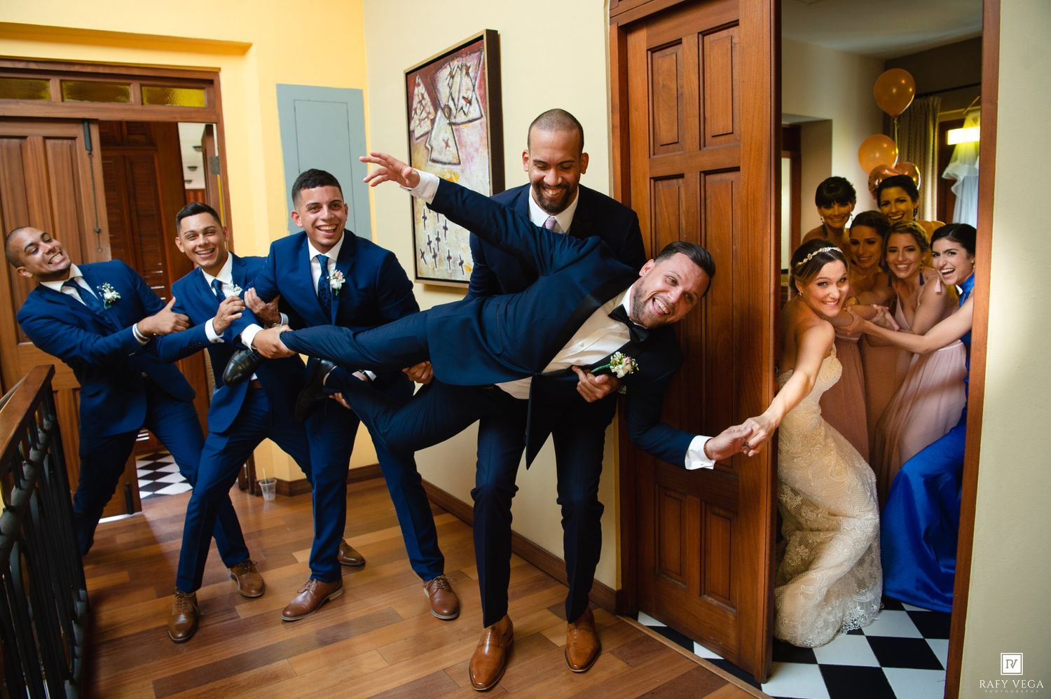 Groom carried by groomsmen in navy suits while bride and bridesmaids laugh in doorway at fun wedding photo.