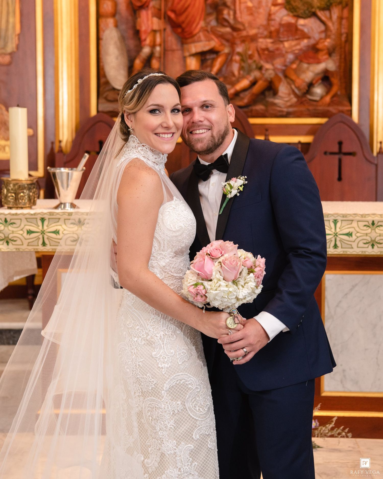 Bride in white lace gown with veil and groom in navy tuxedo pose together holding pink bouquet inside a church.