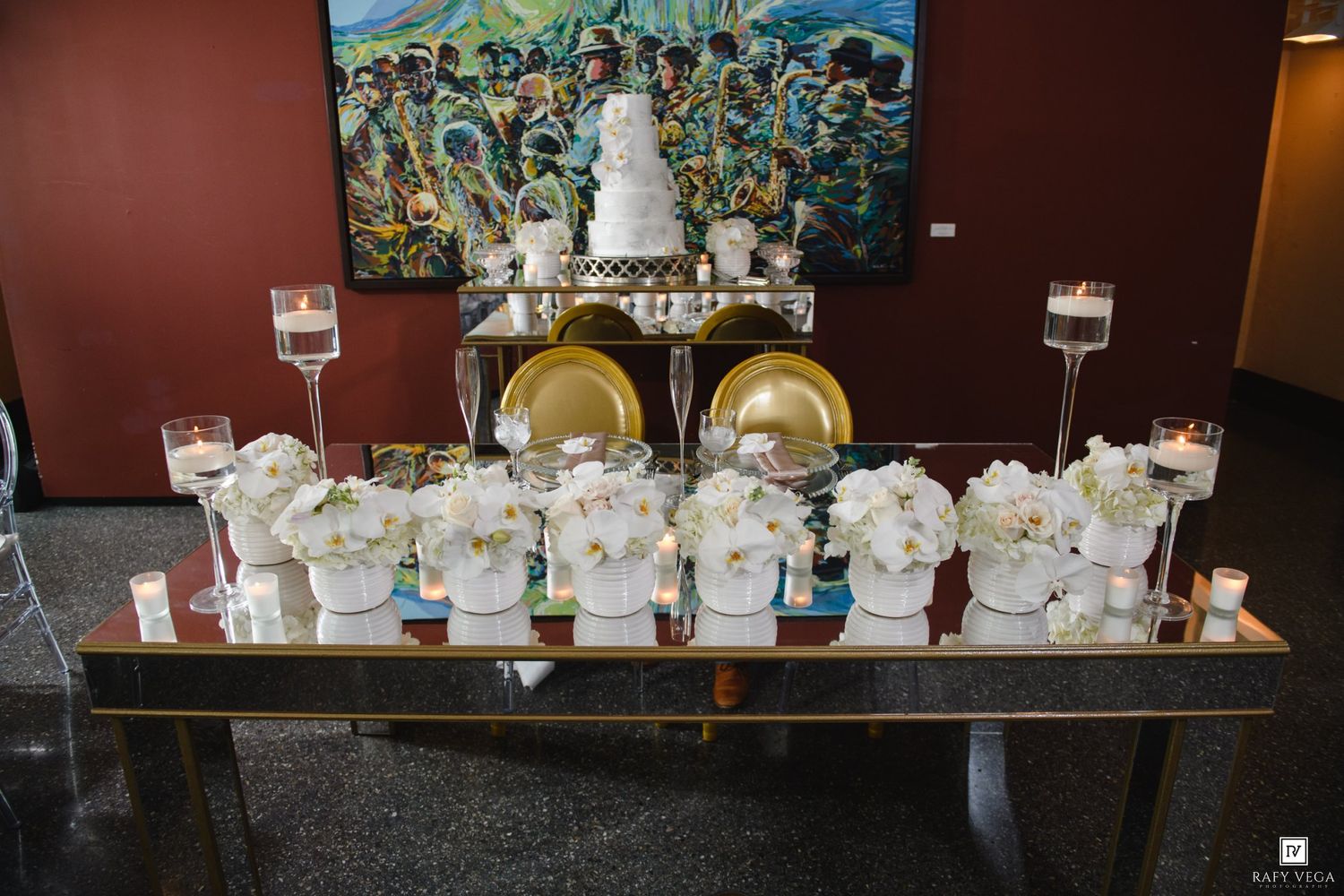 Elegant wedding dessert table with white floral arrangements, gold chargers, candles, and tiered cake against colorful artwork.