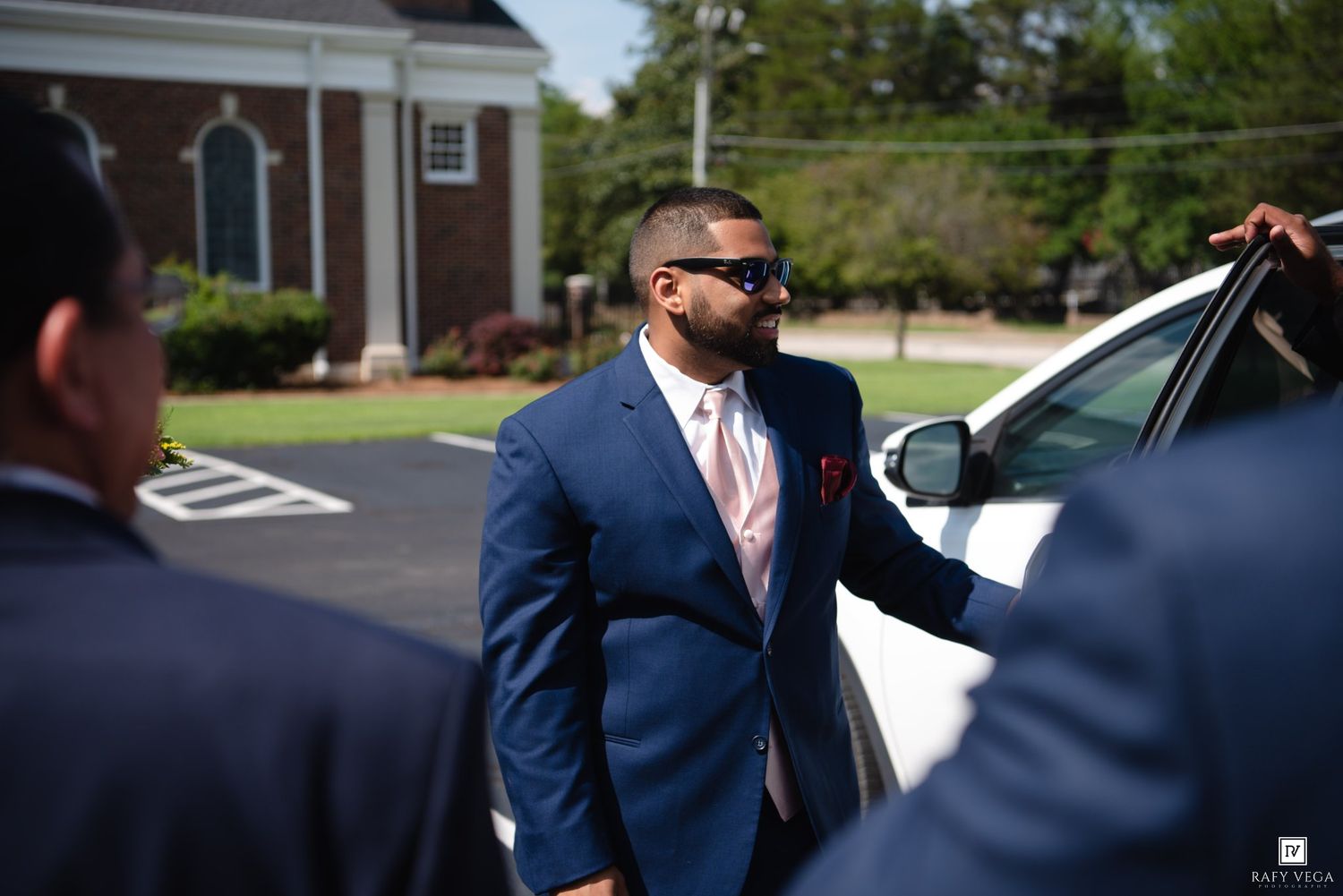 Man in blue suit and sunglasses exits white car outside brick building on sunny day.