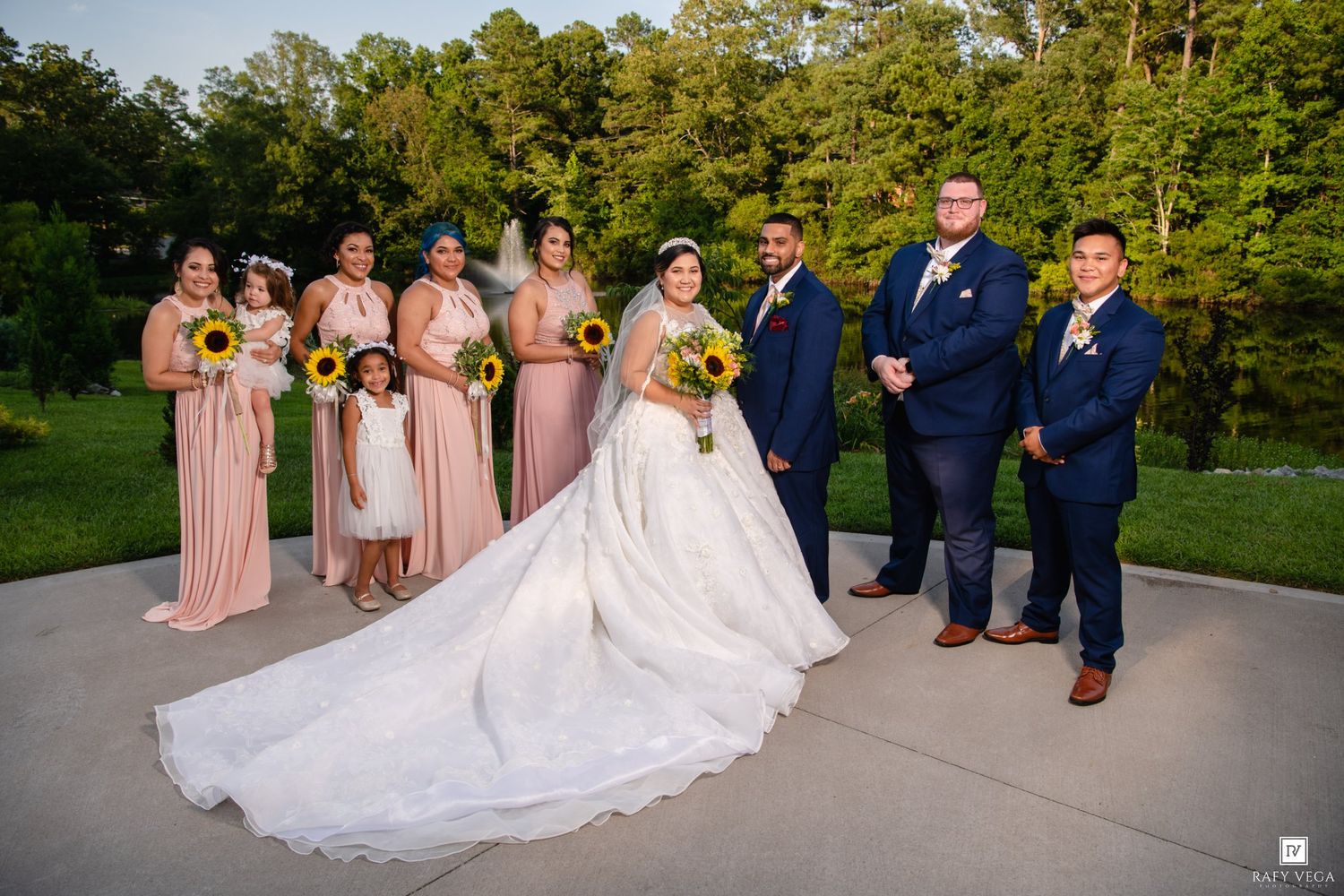 Wedding party poses outdoors, bride in white gown with train, bridesmaids in blush pink, groomsmen in navy suits, sunflower bouquets.