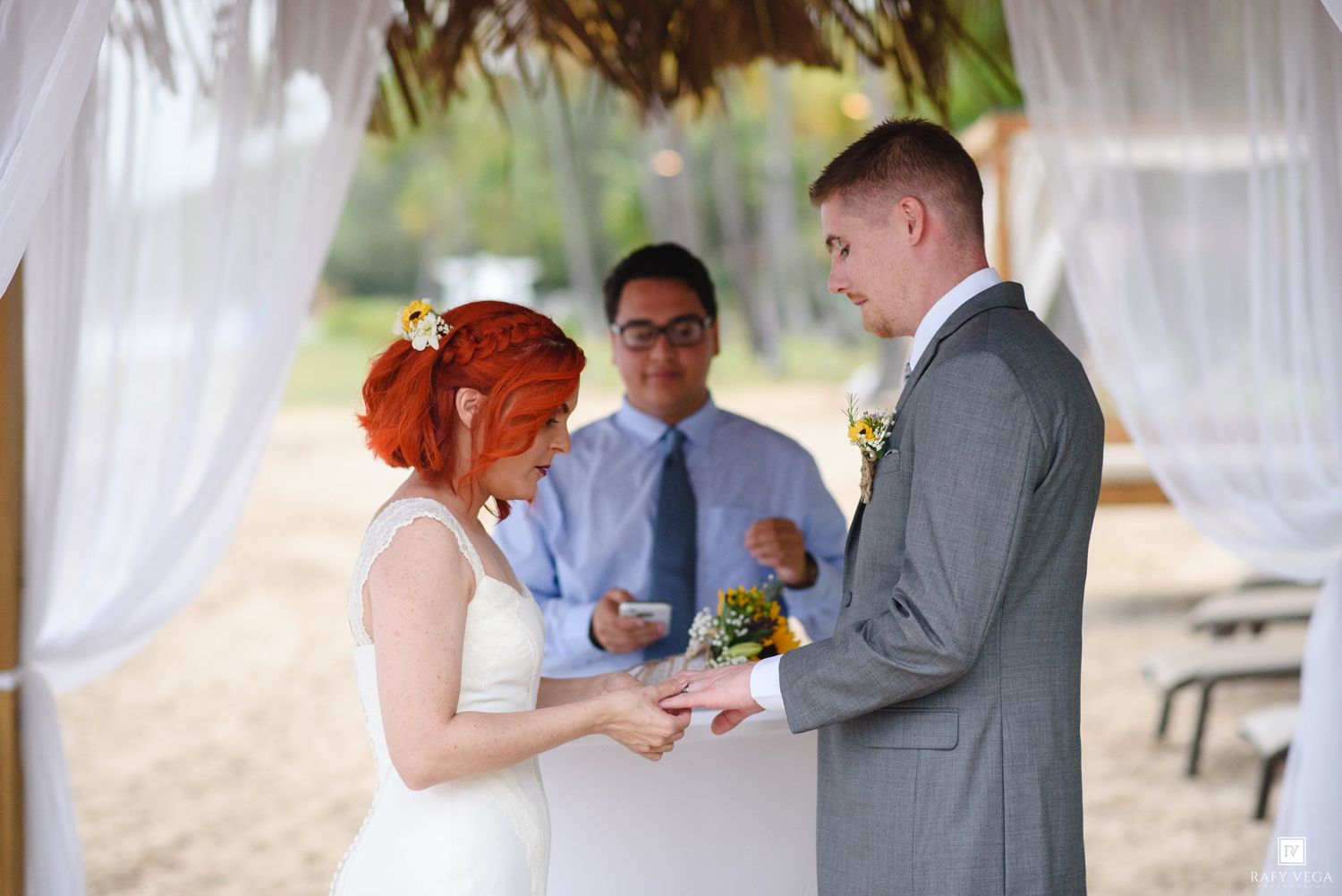 Bride placing wedding ring on groom's finger during outdoor ceremony with officiant and white draping.