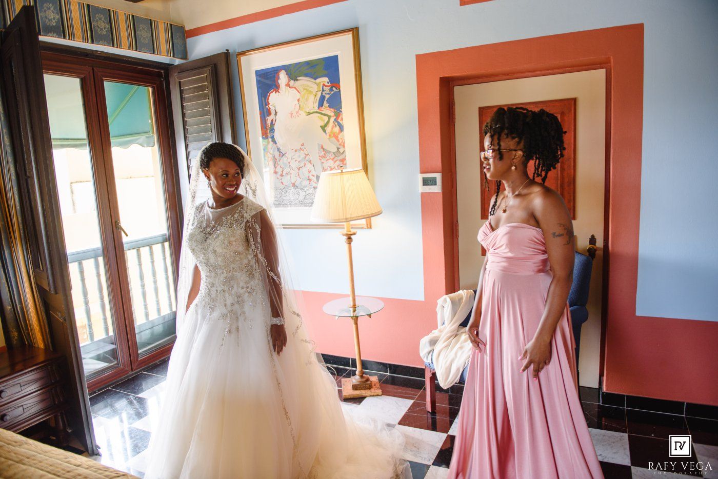 Bride in white gown with cape and bridesmaid in pink dress stand in elegant room during wedding preparations.