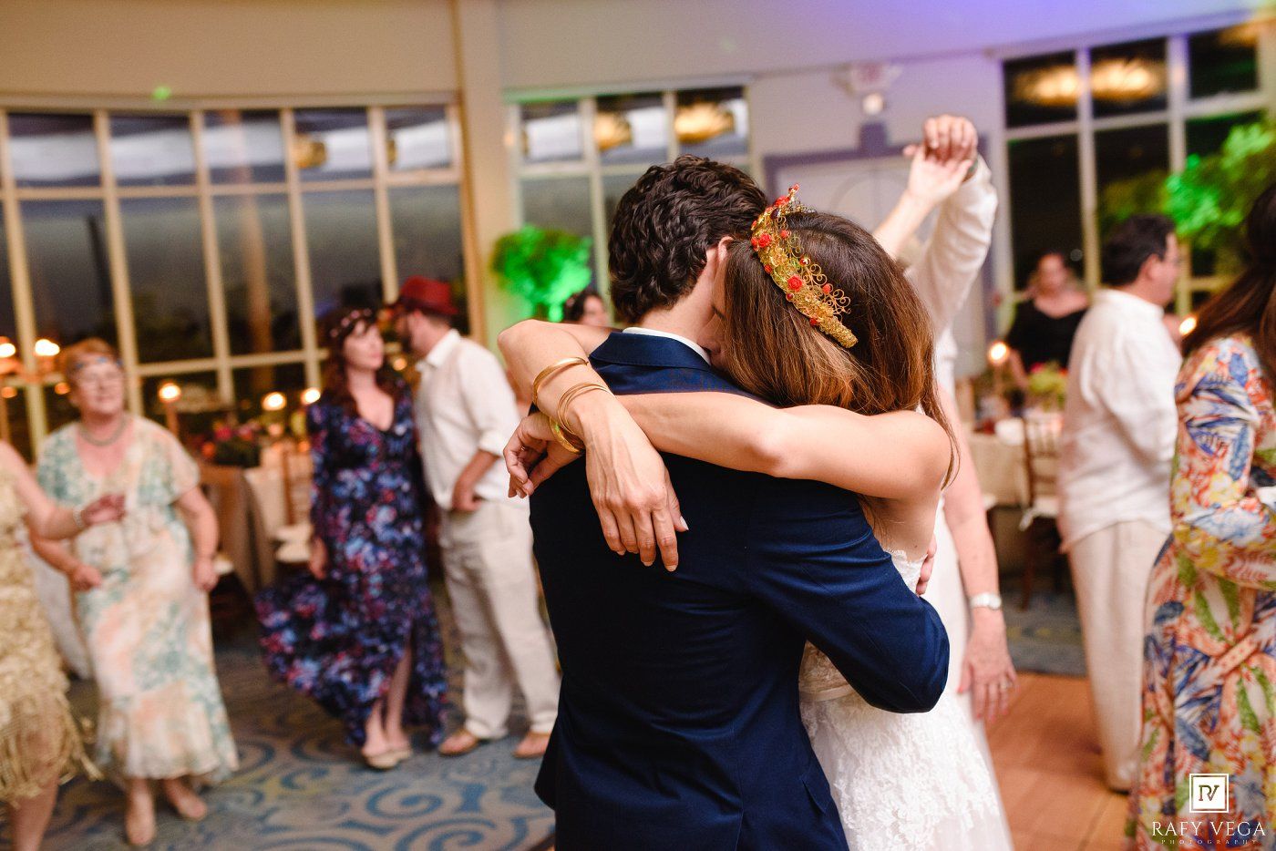 Bride and groom share an emotional first dance embrace at elegant wedding reception with guests dancing in background.