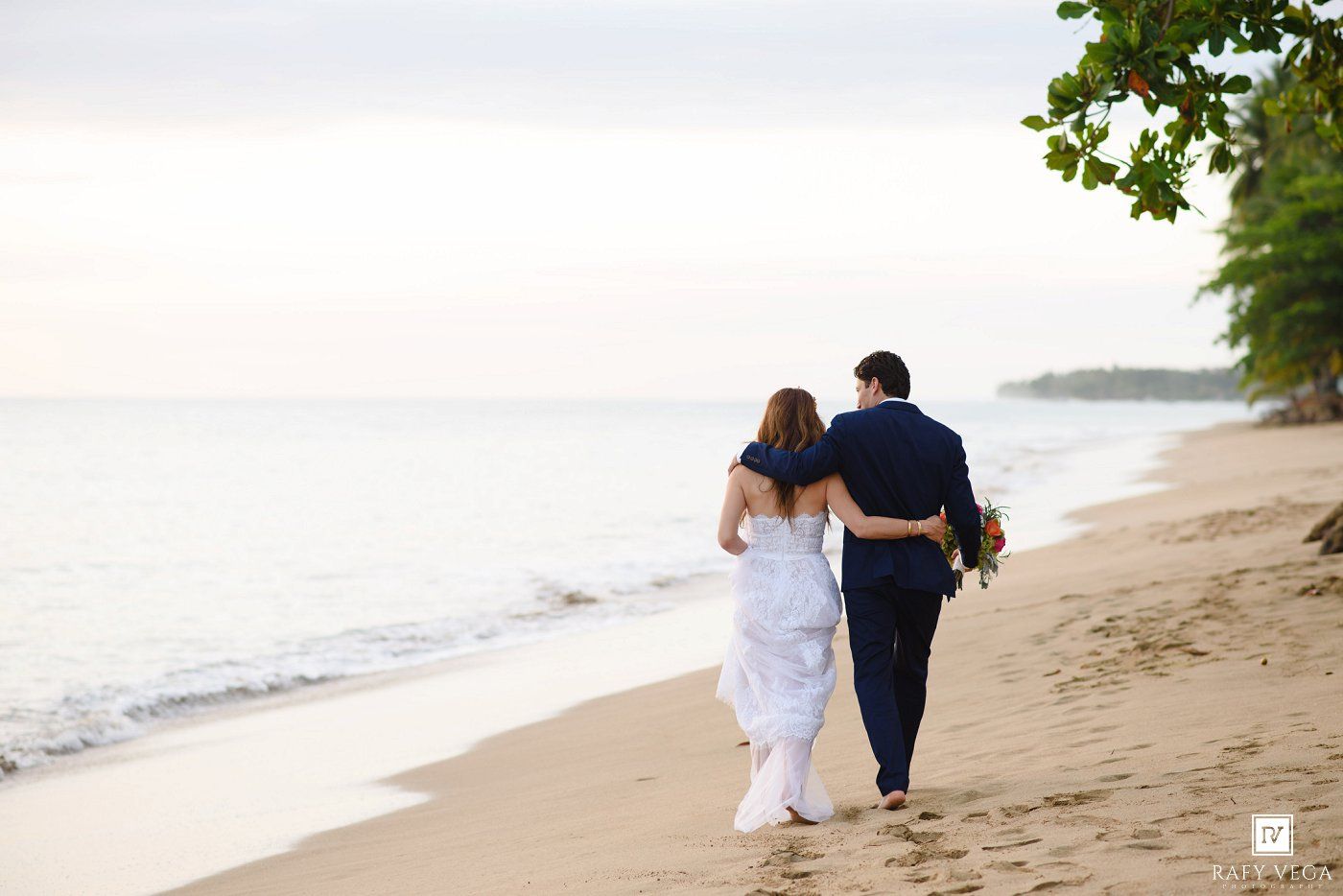 Newlywed couple walks hand in hand along a tropical beach at sunset after their wedding.