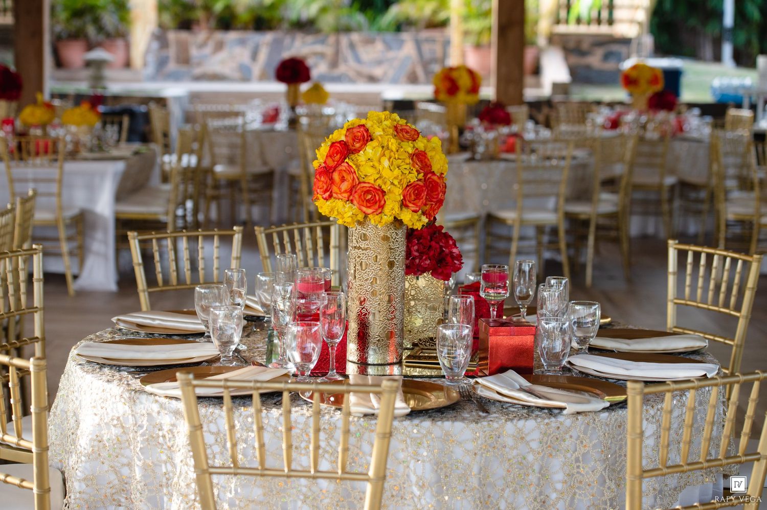 Elegant wedding reception tables with gold chiavari chairs, red and yellow floral centerpieces, and white lace tablecloths.
