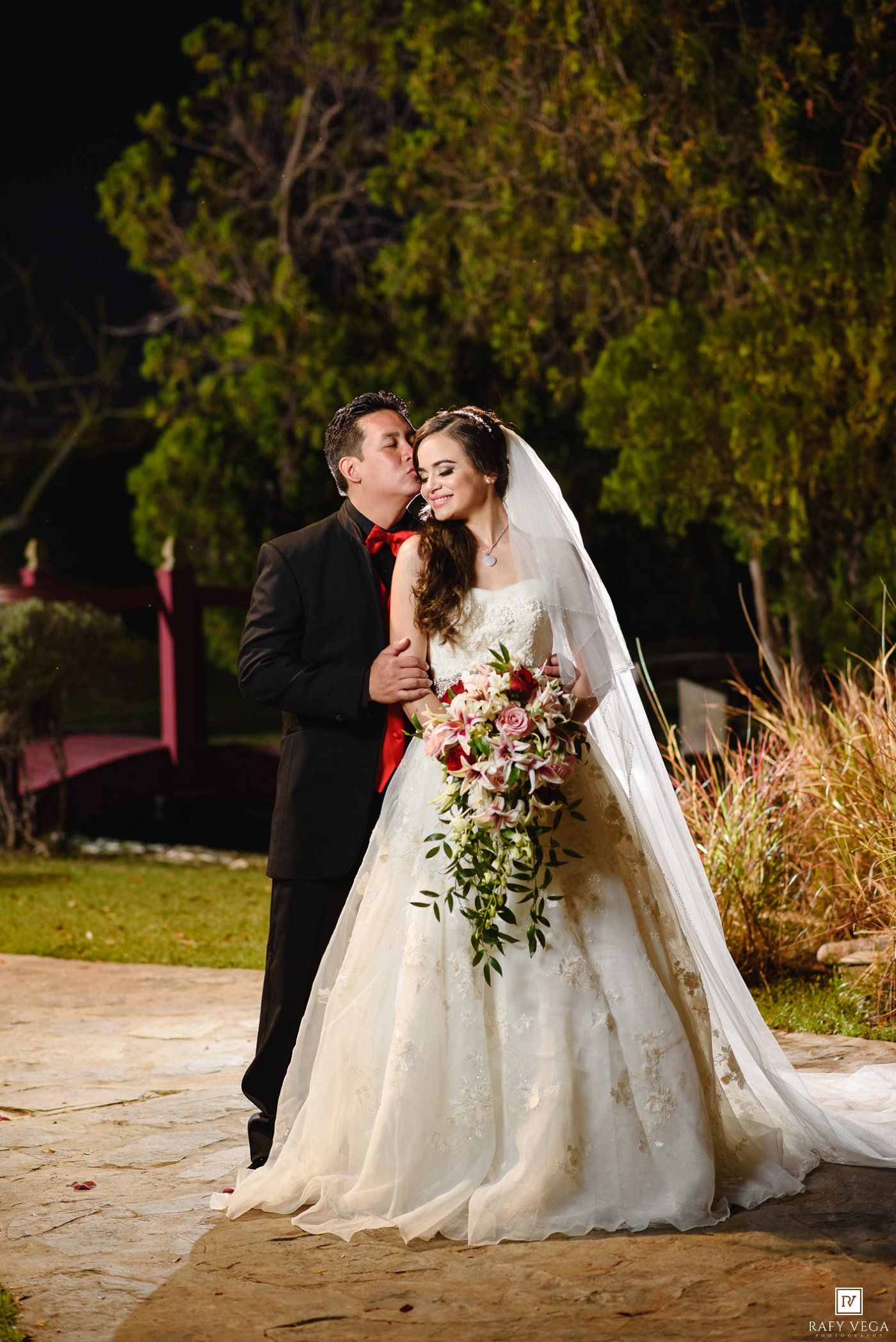 Groom in black tuxedo kisses bride in white lace gown holding floral bouquet during outdoor wedding night ceremony.