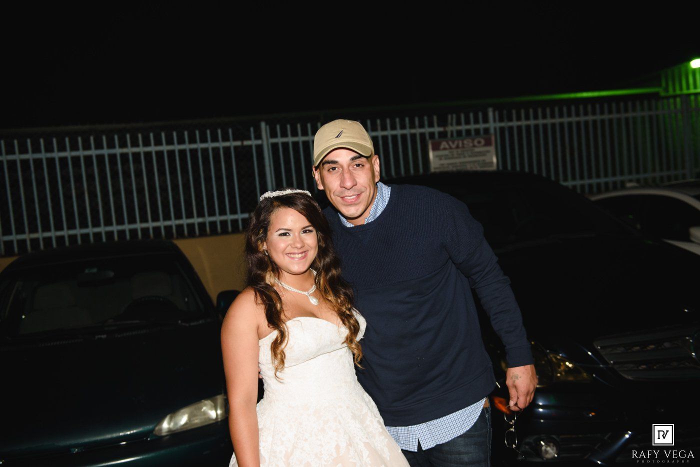 Bride in white gown poses with man in blue sweater and cap outside at night near parked cars.