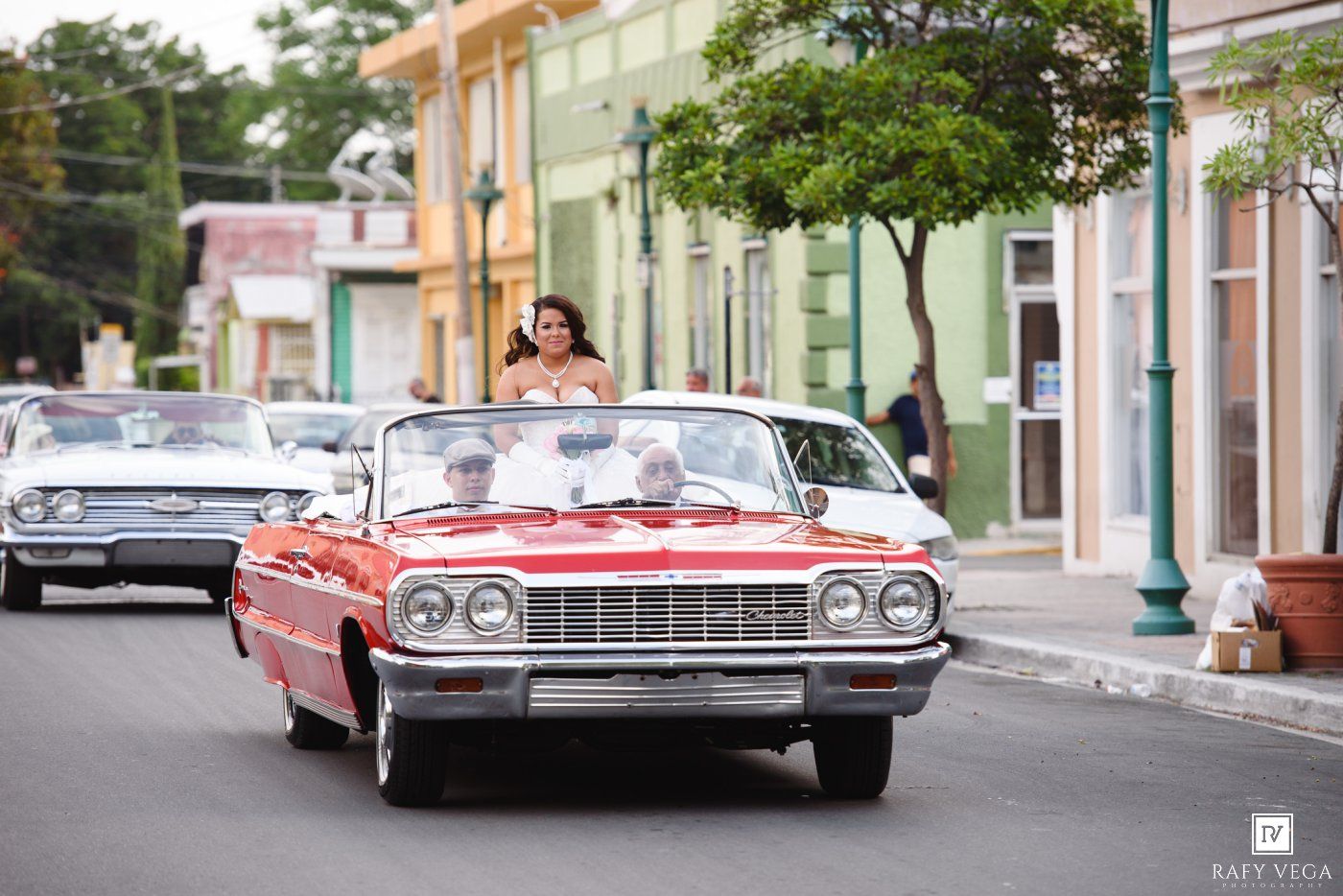 Bride rides in a classic red convertible car through a colorful street during a wedding procession.