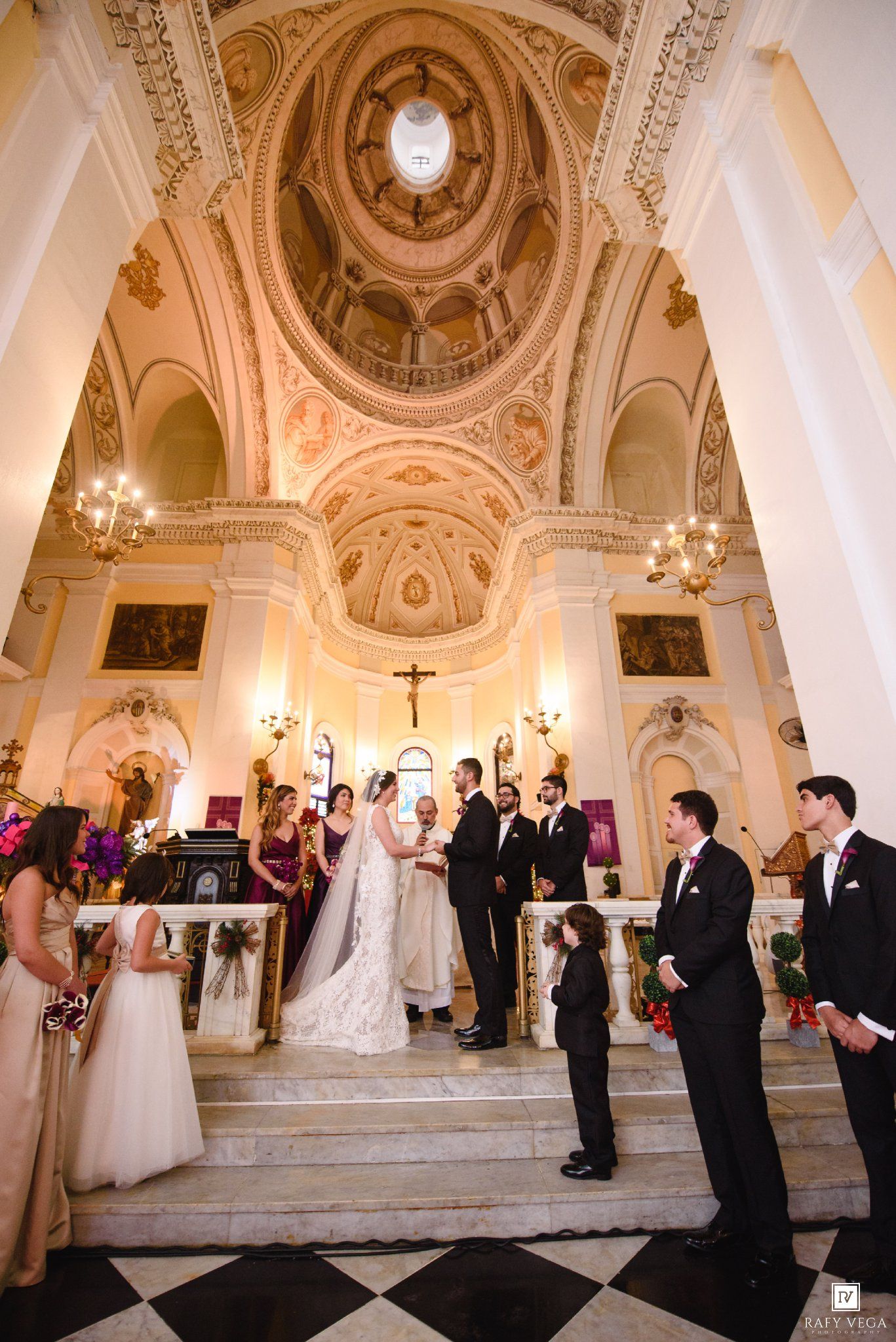 Elegant wedding ceremony in a grand baroque church with ornate domed ceiling, bride and groom at altar with wedding party.