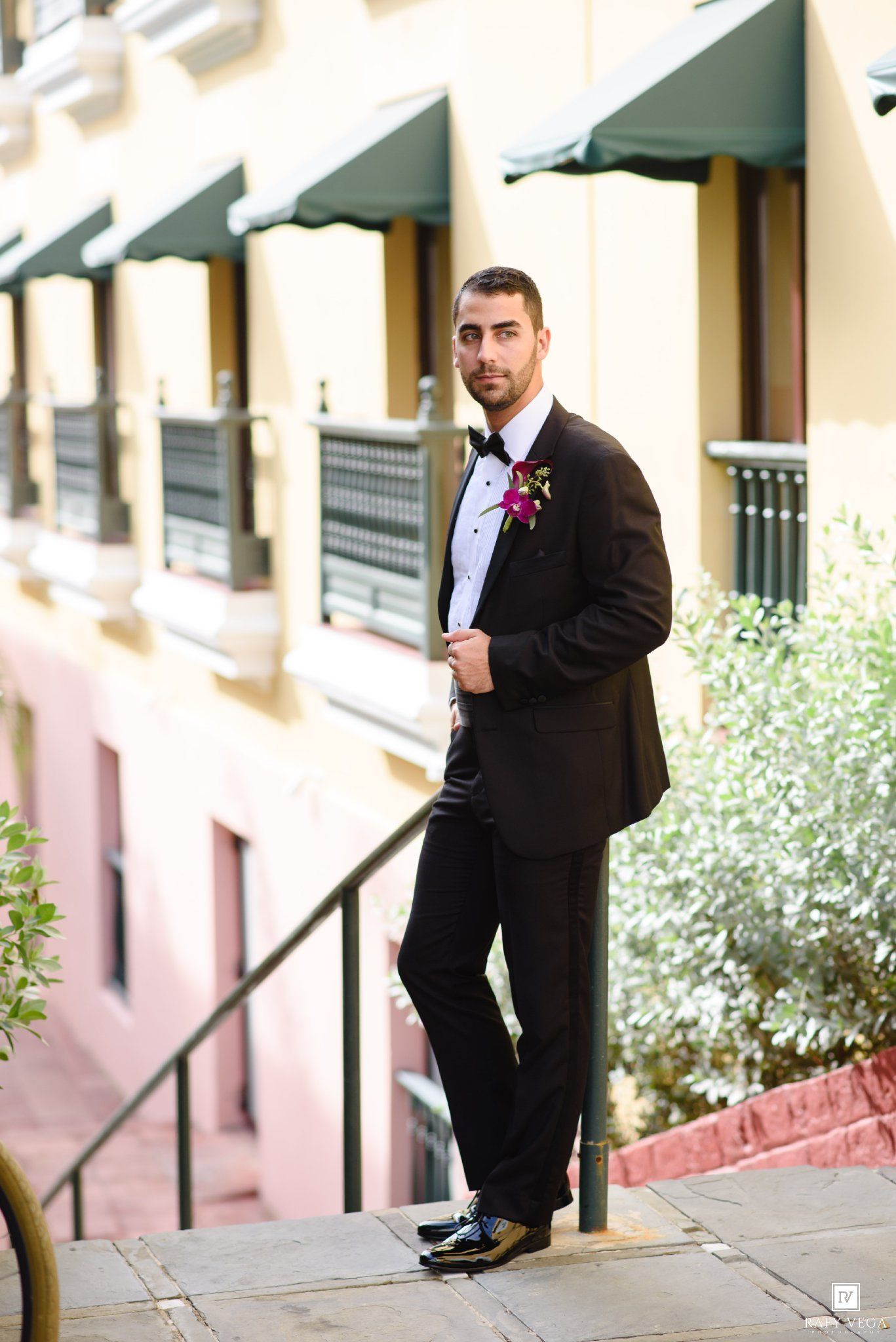 Groom in black tuxedo with bow tie and boutonniere posing on steps outside elegant building.