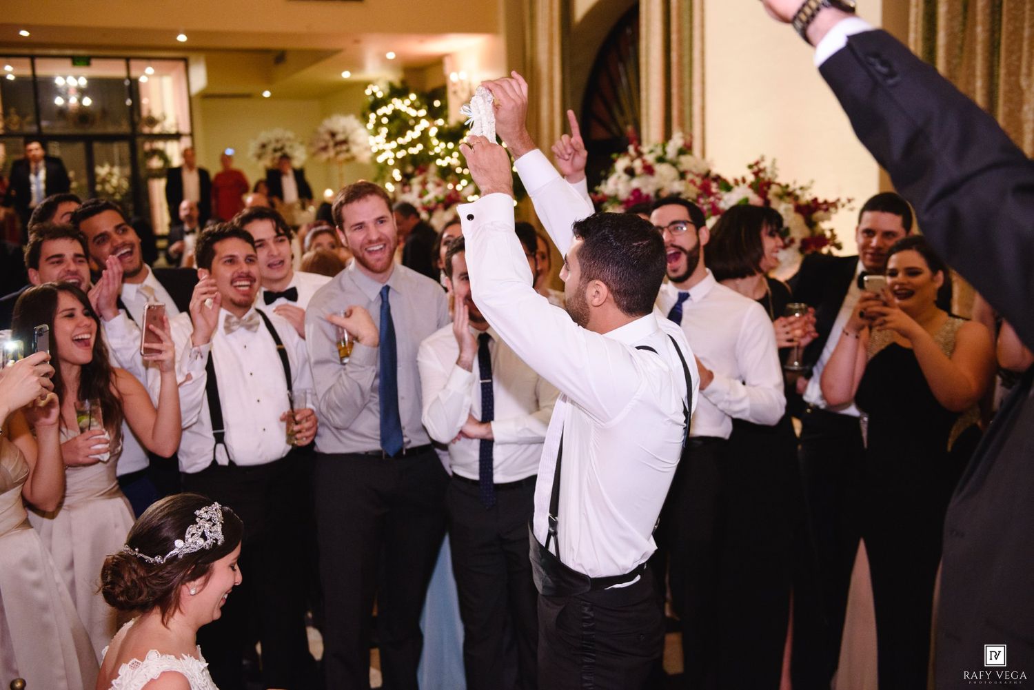 Groom celebrates with groomsmen in suspenders at elegant wedding reception, guests cheering with raised arms.