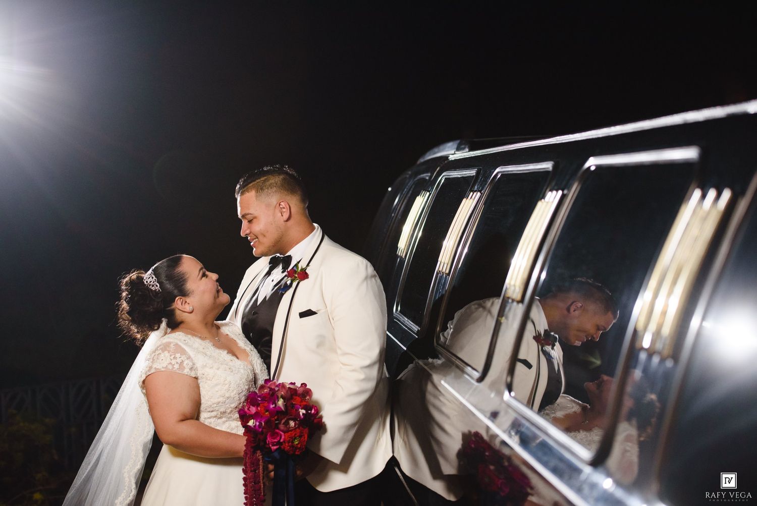 Bride and groom share a romantic moment beside a limousine at night, bride holding a red bouquet in wedding attire.