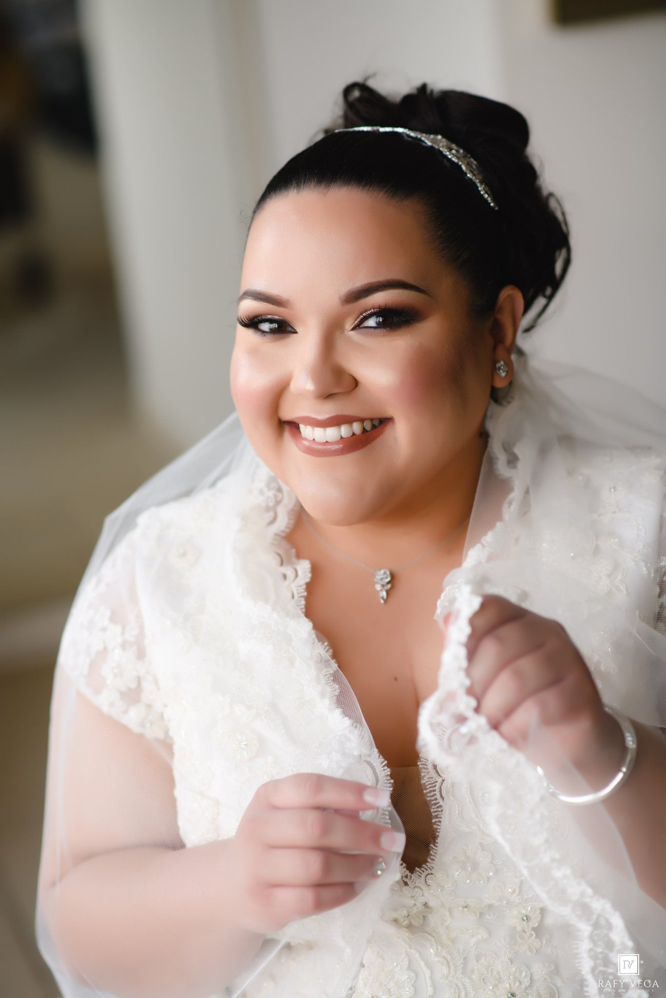 Smiling bride in white lace wedding dress with veil and tiara, wearing elegant jewelry on her special day.