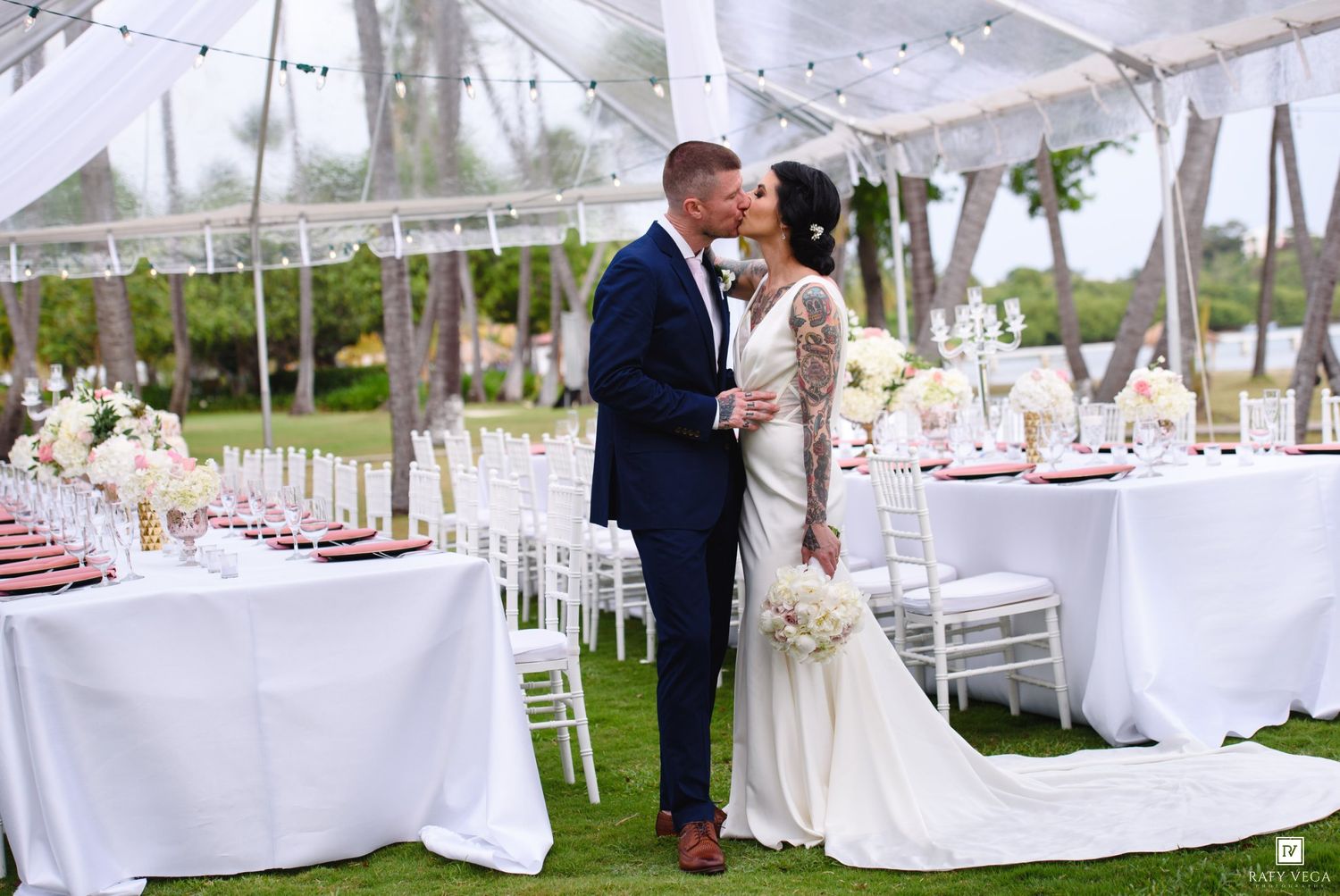 Bride and groom share a kiss under an elegant white wedding tent with floral centerpieces and Chiavari chairs.