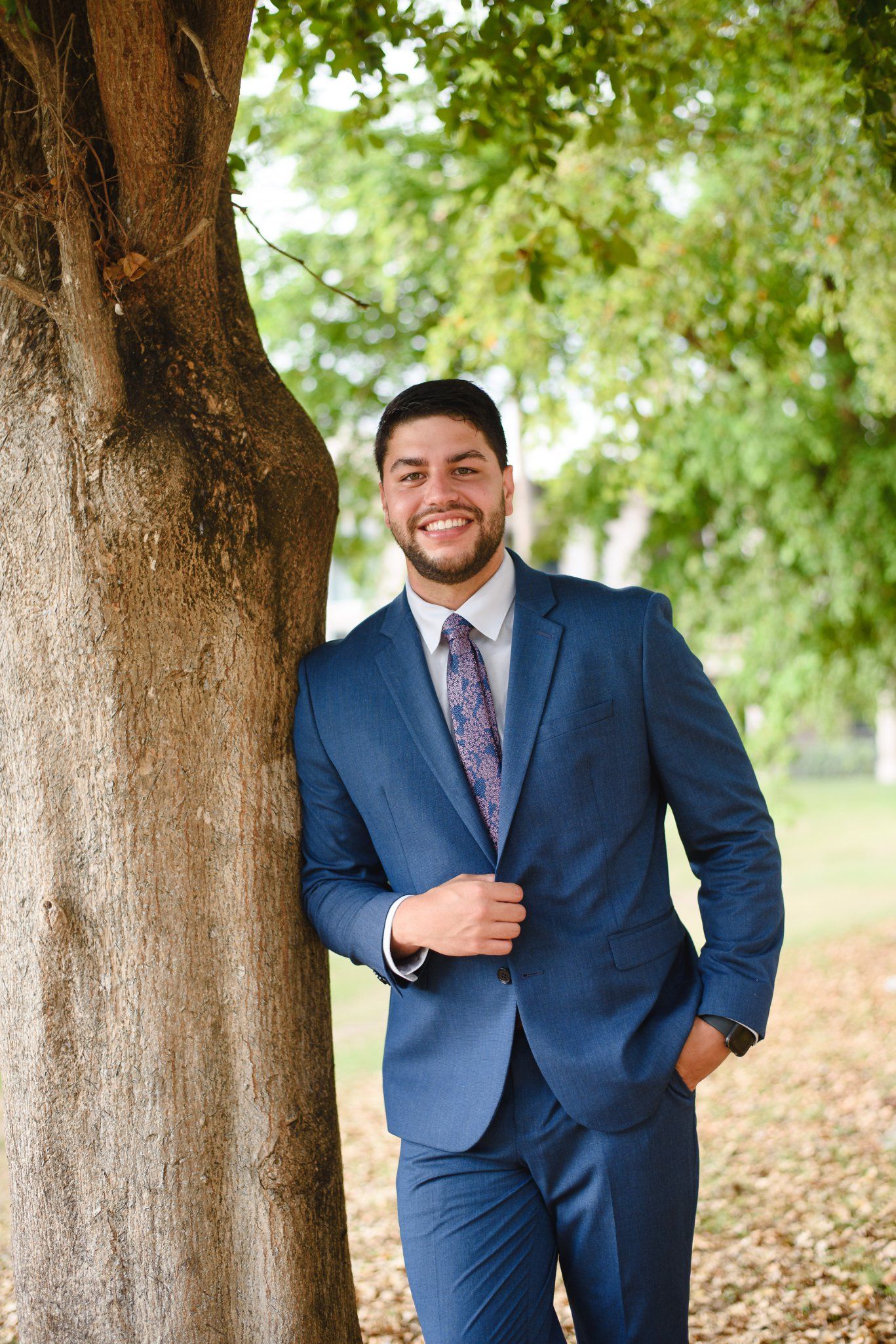 Smiling groom in blue suit and floral tie leans against tree in park.
