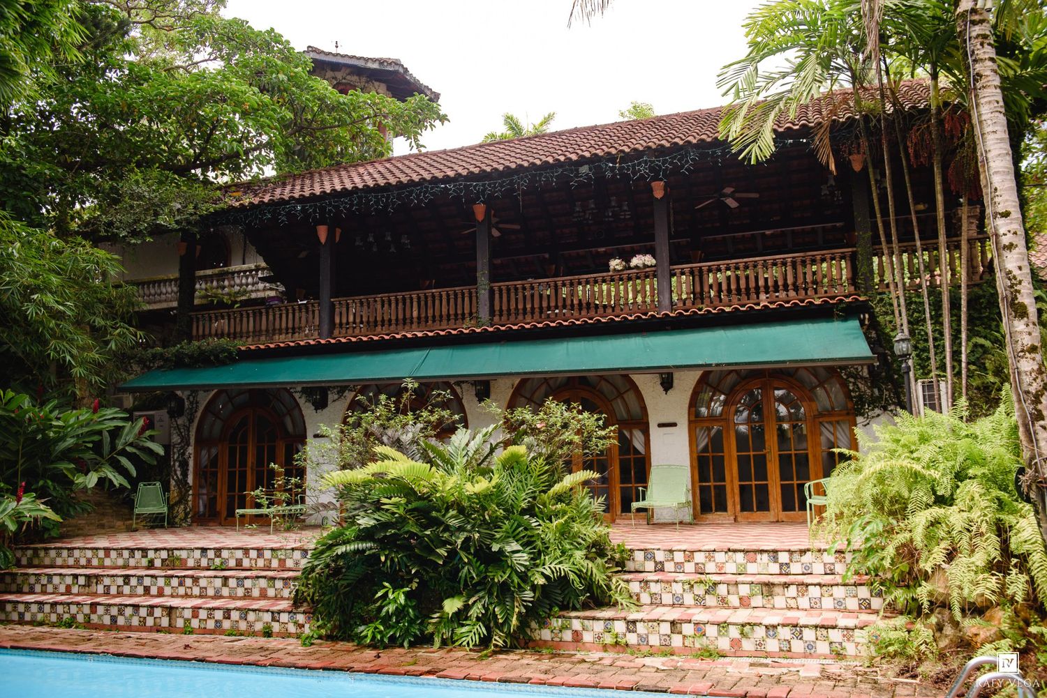 Elegant two-story tropical villa with wooden balcony, arched windows, lush greenery, and a pool in a rainforest setting.