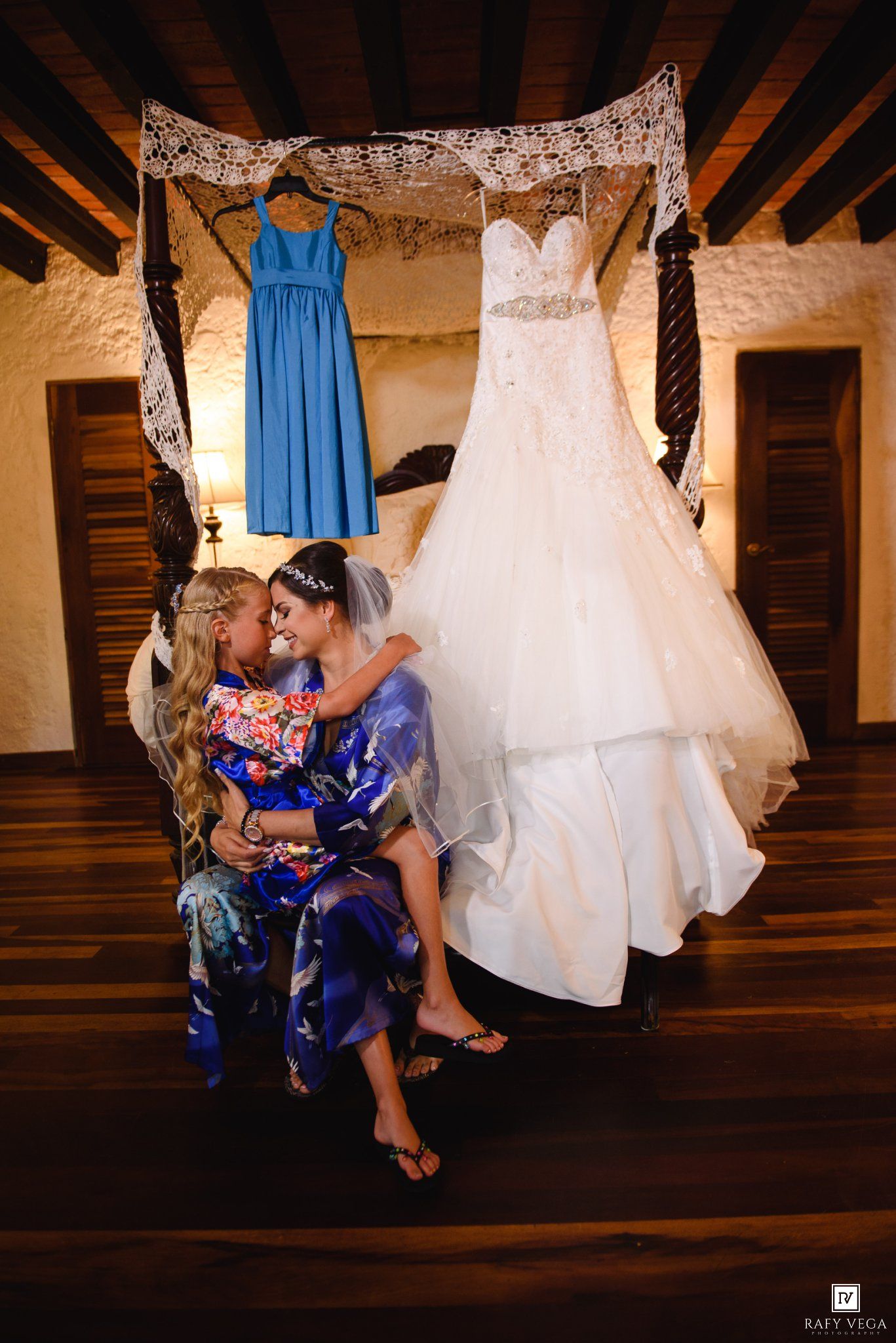 Bride and child share tender moment in robes before wedding, with white gown and blue bridesmaid dress hanging behind them.