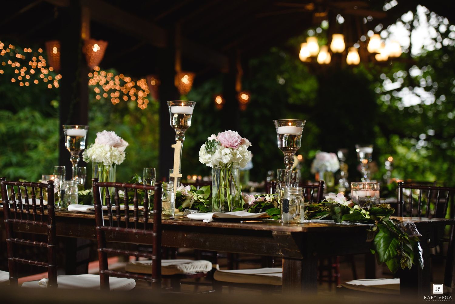 Elegant outdoor wedding reception table with floral centerpieces, candles, and string lights in a lush garden setting.