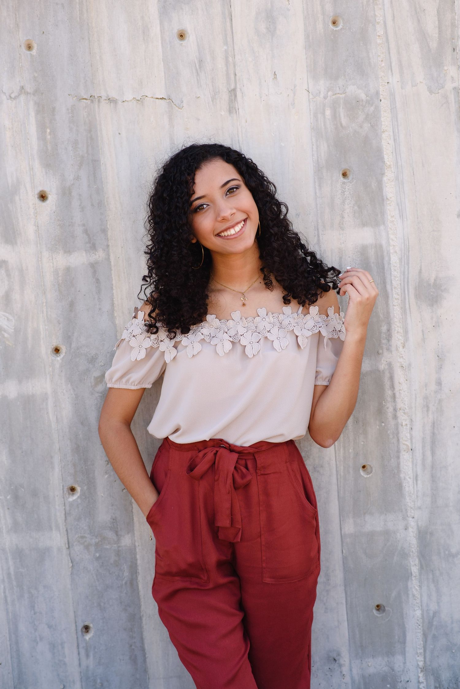 Smiling young woman with curly hair wearing off-shoulder lace top and red pants posing against concrete wall.
