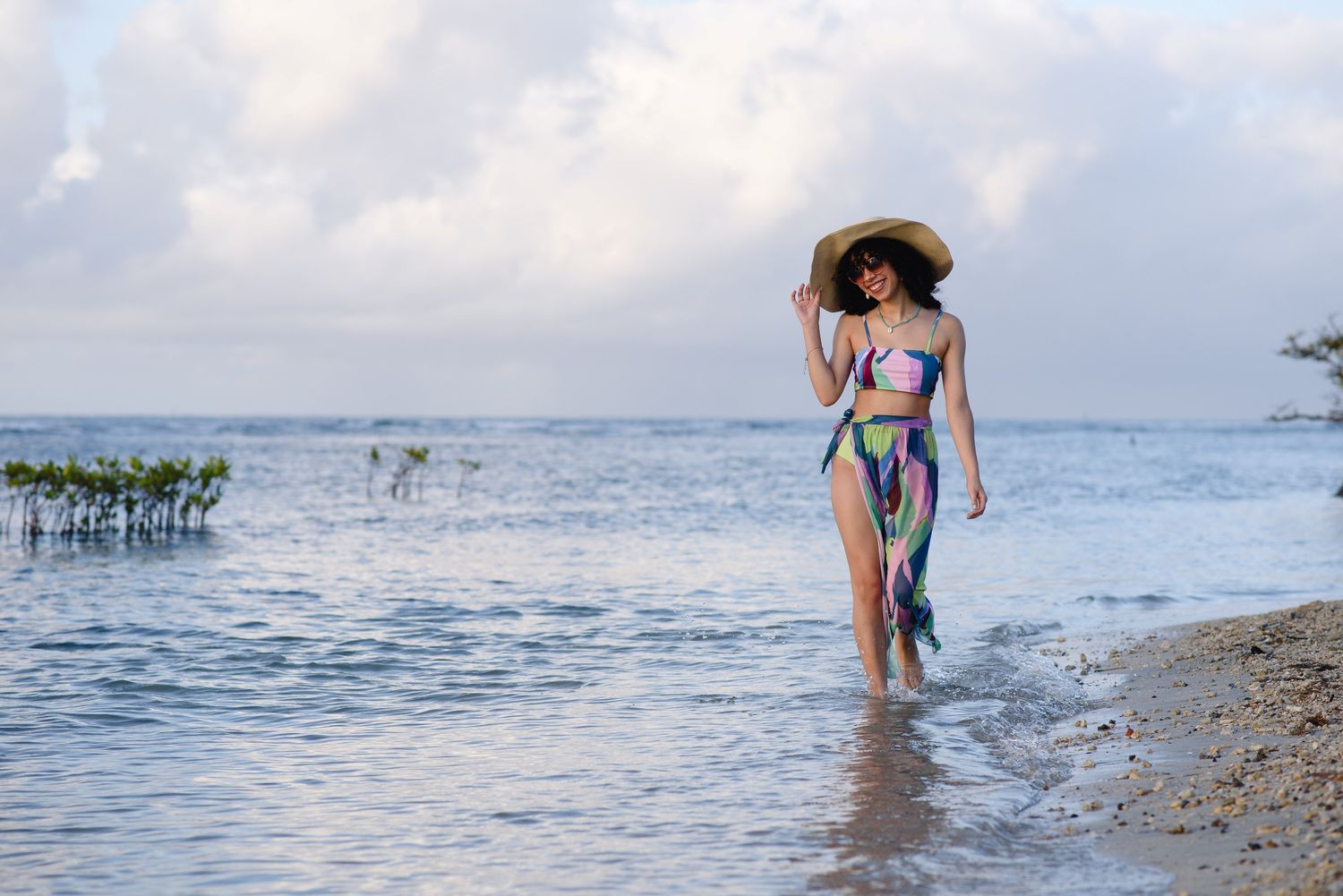 Woman in colorful bikini and straw hat walking along a tropical beach shoreline with calm waters.