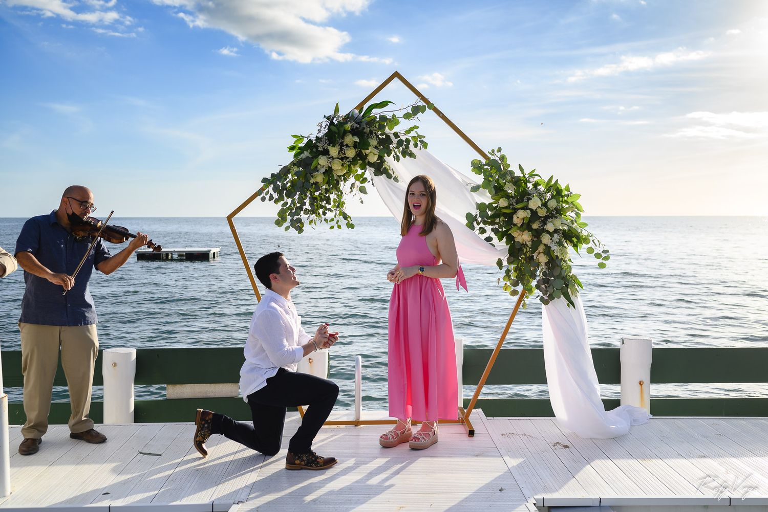 Man proposes on a decorated pier with floral arch, violinist, and ocean backdrop, as woman in pink dress smiles.