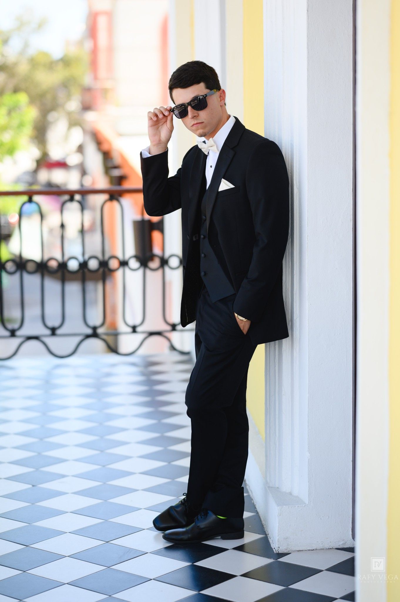 Young man in black tuxedo and sunglasses leaning against white column on checkered floor balcony.