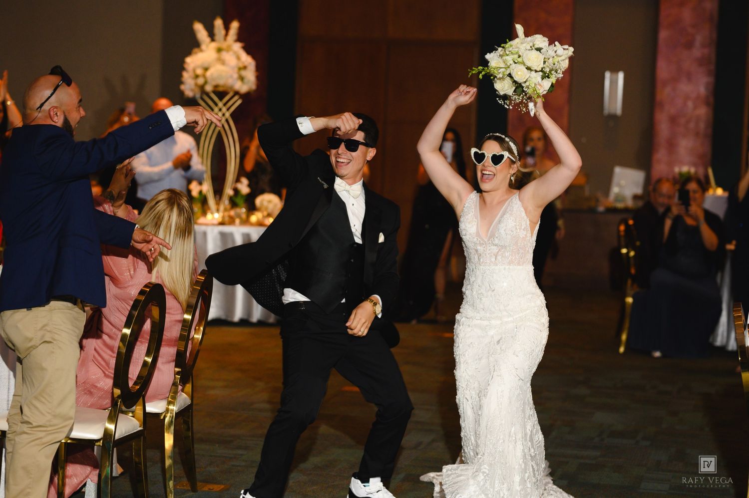 Bride and groom dancing joyfully at wedding reception, bride holding bouquet aloft wearing sunglasses, groom in tuxedo.