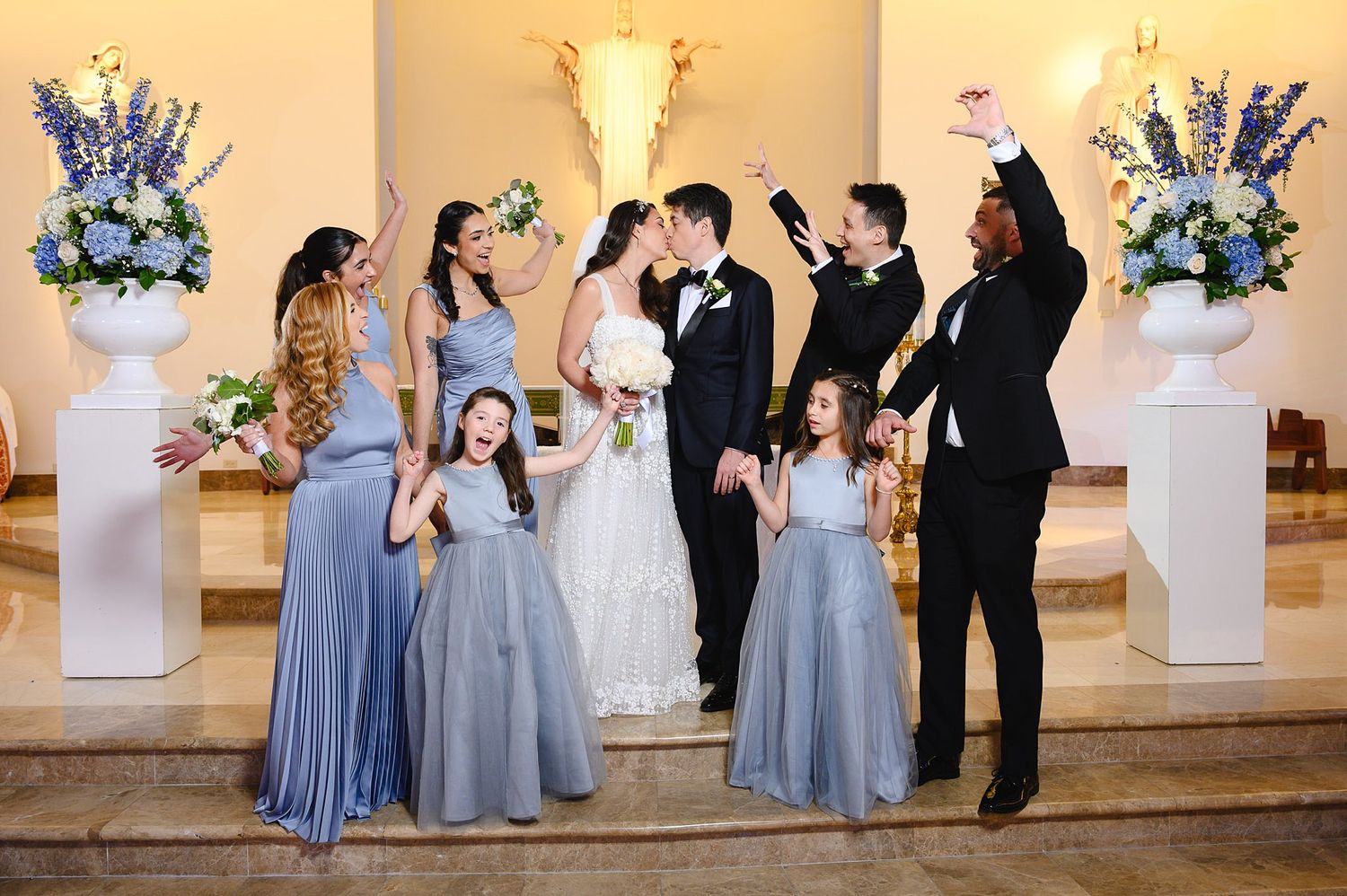 Wedding party celebrates in church, bride and groom kissing, bridesmaids in blue gowns, groomsmen cheering, flower girls smiling.