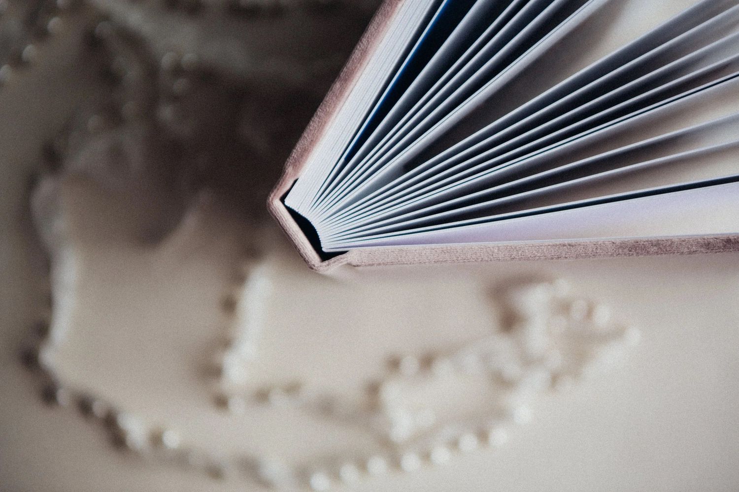 Close-up of an open hardcover book with white pages fanned out, beside a delicate pearl necklace on a beige surface.