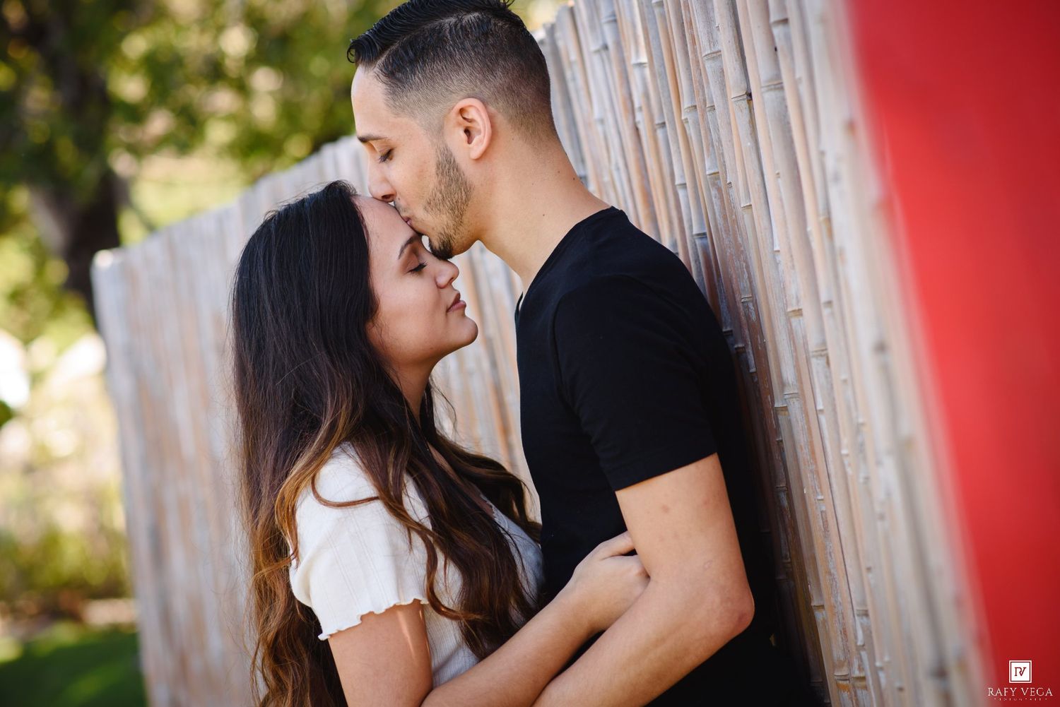Couple sharing a tender kiss against a wooden fence outdoors, surrounded by lush greenery in a romantic engagement photo.