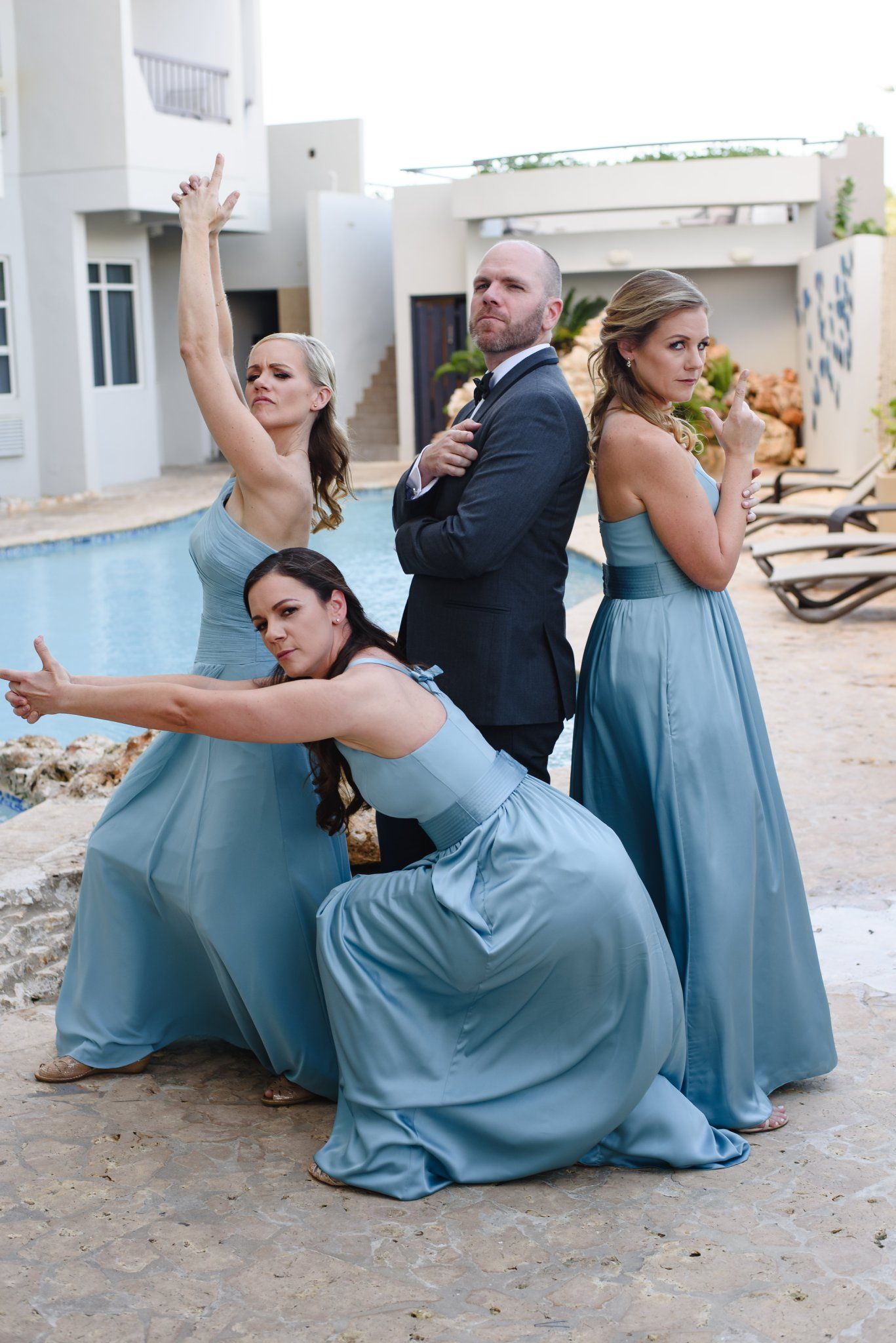 Bridesmaids in blue gowns pose dramatically with a suited groomsman poolside at a destination wedding.