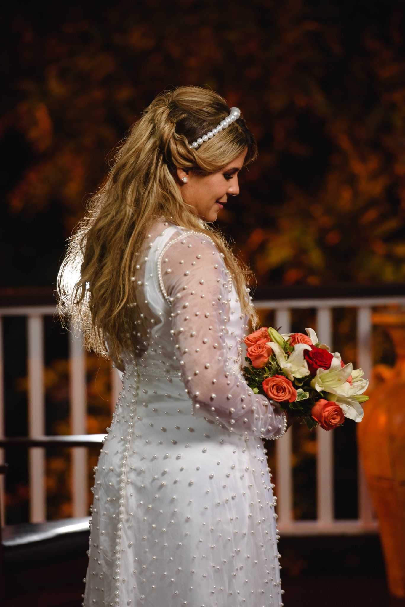 Bride in white polka dot long-sleeve gown holding red and white bouquet, smiling downward with pearl headband.